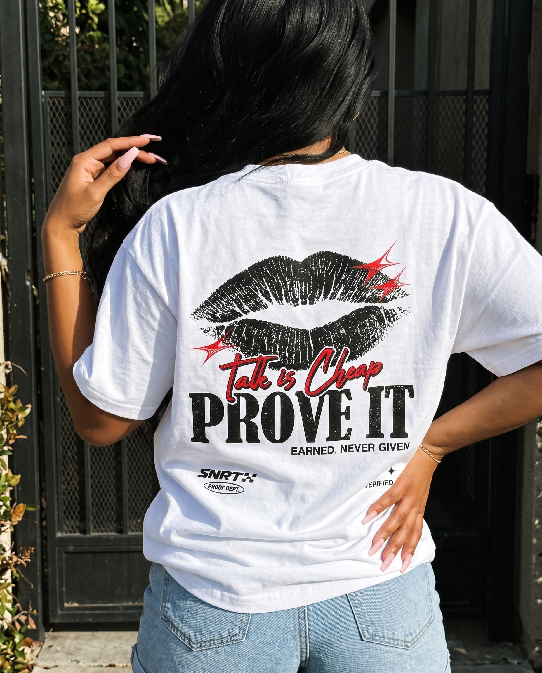 A woman viewed from behind wearing a white graphic T-shirt with a large black lip print, red and black text reading 'Talk is Cheap PROVE IT', and light blue jeans, standing outdoors in front of a dark metal gate.