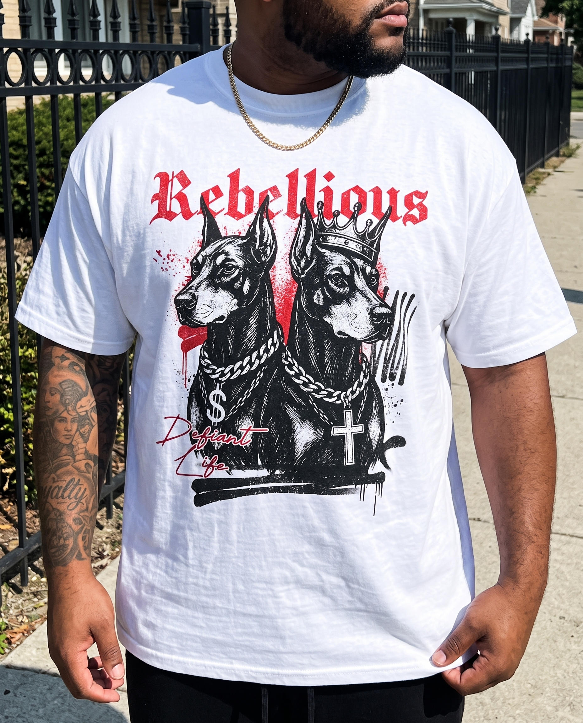 A man wearing a white graphic t-shirt featuring two black dogs wearing crowns and chains, with the word 'Rebellious' in red gothic lettering above them. The man is standing outdoors on a sidewalk next to a black metal fence.