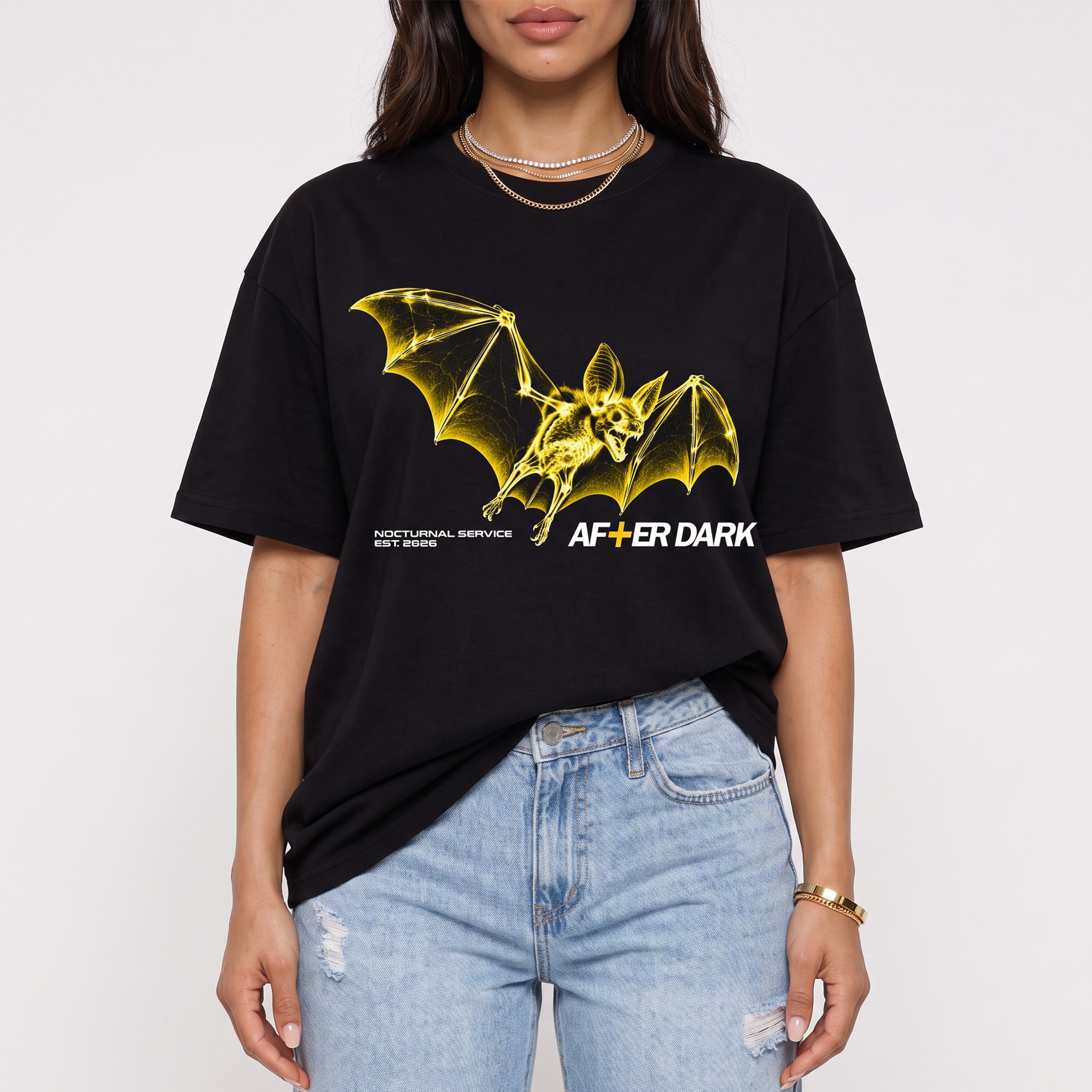 A woman wearing a black graphic t-shirt with a yellow bat design and the text 'AFTER DARK' and 'MOTIONAL SERVICE EST. 2026', paired with light blue jeans, against a plain white background.