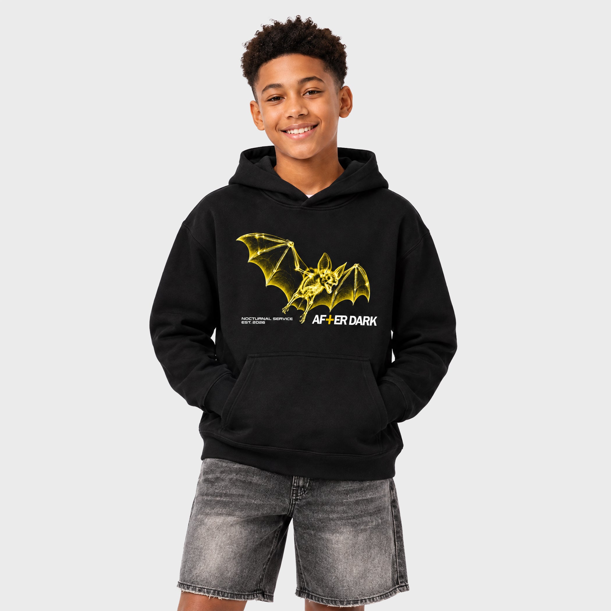 A smiling young man wearing a black pullover hoodie with a yellow bat graphic and the text 'AF+ER DARK' on the front, paired with gray denim shorts, against a light gray studio background.