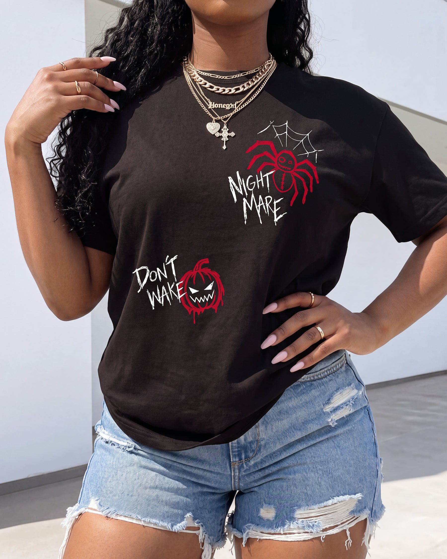 A woman wearing a black graphic t-shirt with red and white spider-themed designs and text, paired with distressed denim shorts.