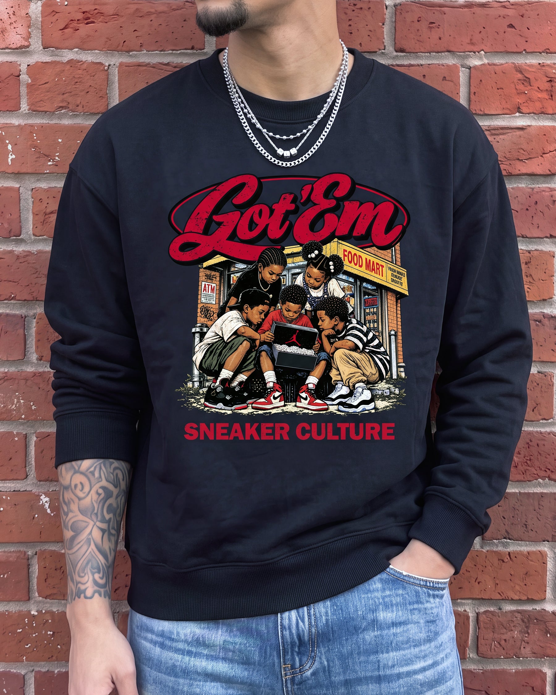 A man wearing a black crewneck sweatshirt with a colorful graphic print of the K-pop group GOT 'EM and the text 'Got 'Em' and 'Sneaker Culture', paired with blue jeans in front of a red brick wall.