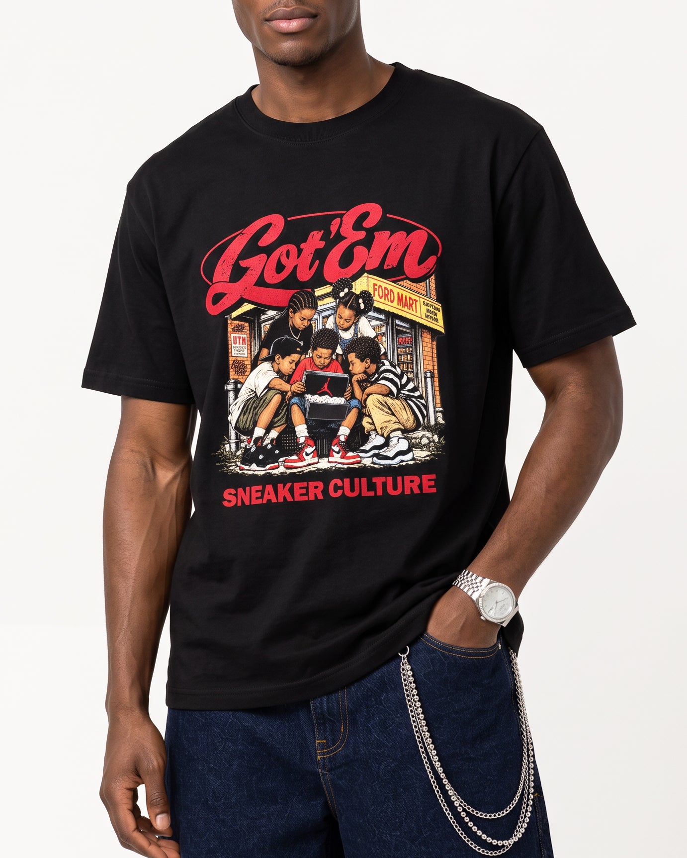 A man wearing a black graphic t-shirt featuring the red and yellow 'Got 'Em Sneaker Culture' print, paired with dark blue jeans and silver chain accessories, against a plain white background.