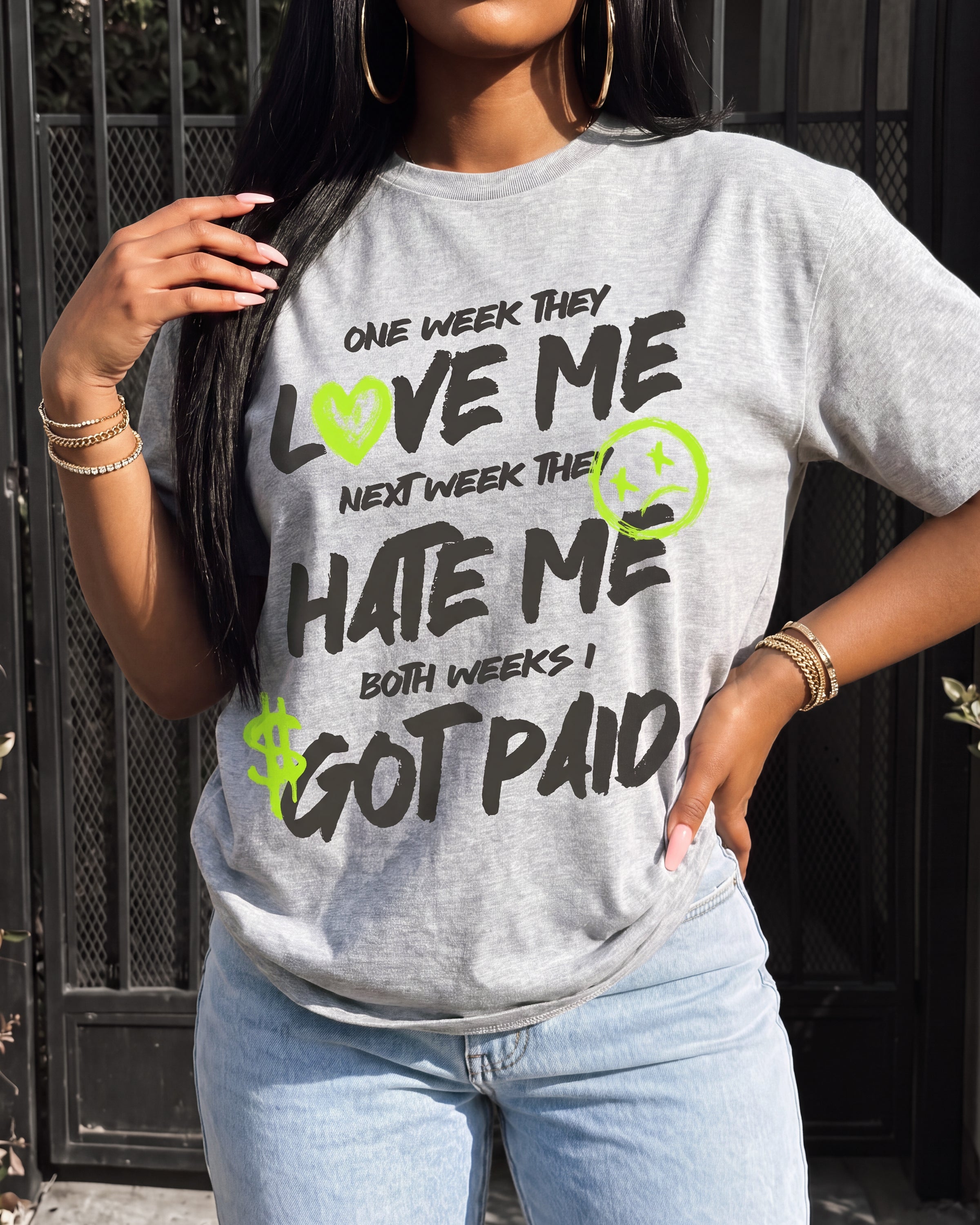 A woman wearing a light heather gray graphic t-shirt with the text 'ONE WEEK THEY LOVE ME NEXT WEEK THEY HATE ME BOTH WEEKS I GOT PAID' in black, accented with lime green smiley and dollar sign graphics. She is wearing light blue jeans and gold jewelry, standing in front of a black metal fence.
