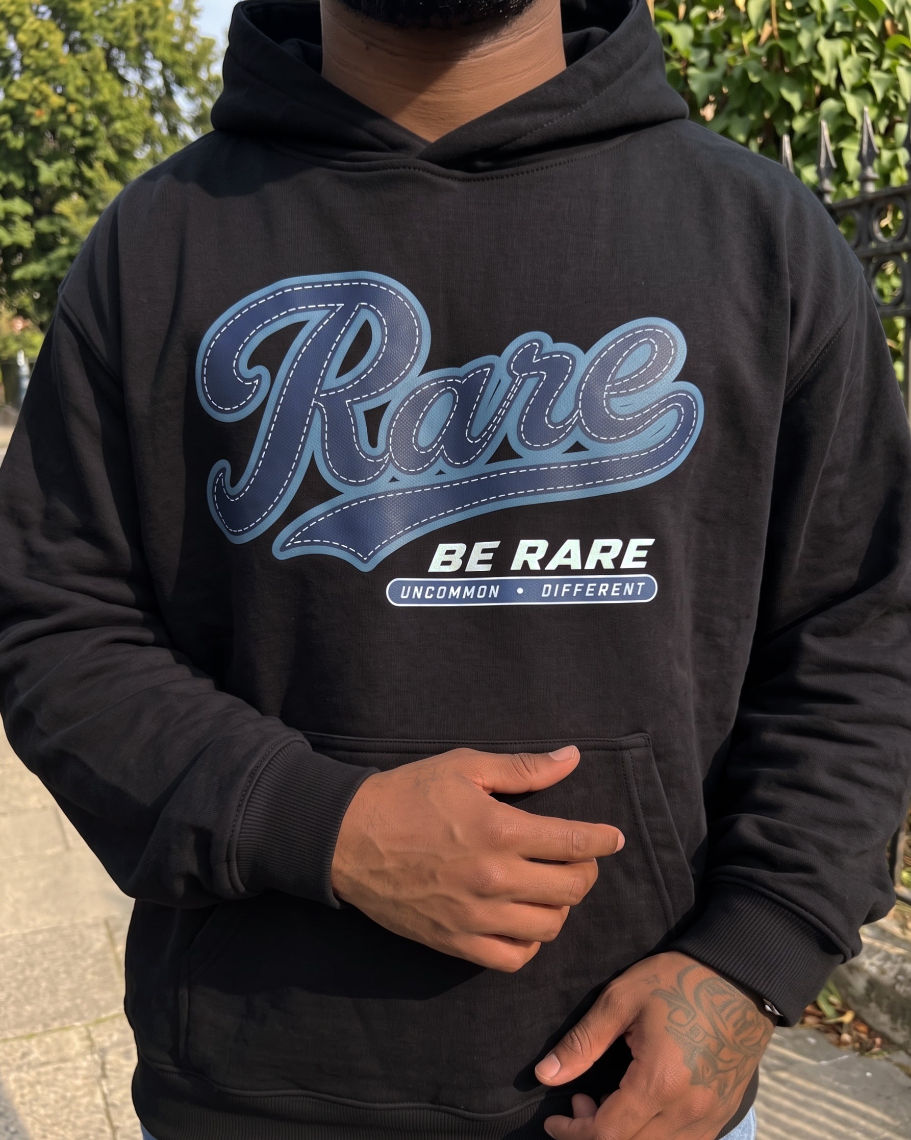 A person wearing a black hoodie with a large blue and white graphic on the chest that reads 'Rare BE RARE' and 'UNCOMMON • DIFFERENT'. The photo is taken outdoors with trees and pavement visible in the background.