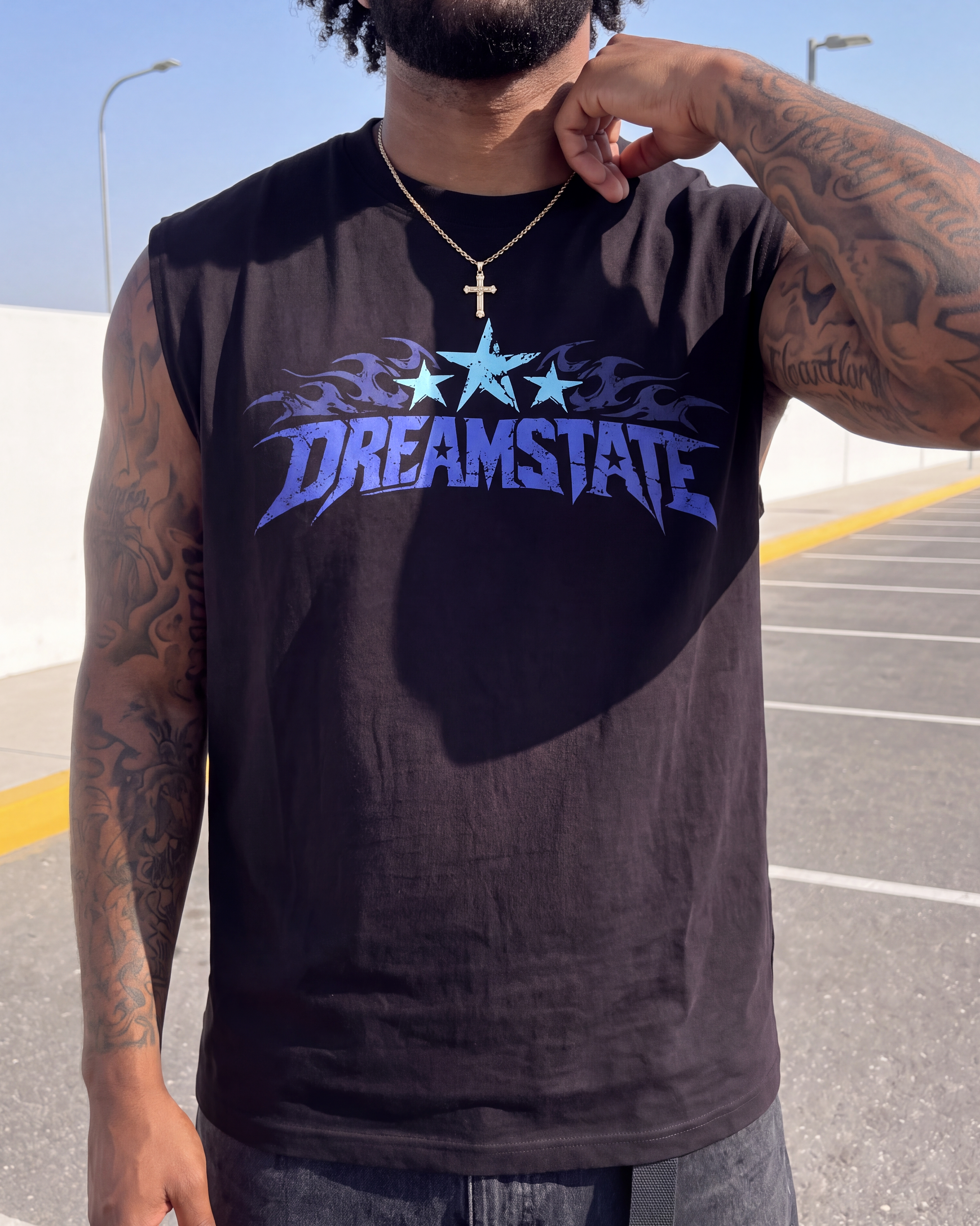 A man wearing a black sleeveless t-shirt with a blue and light blue 'DREAMSTATE' graphic across the chest, standing in an outdoor parking lot.