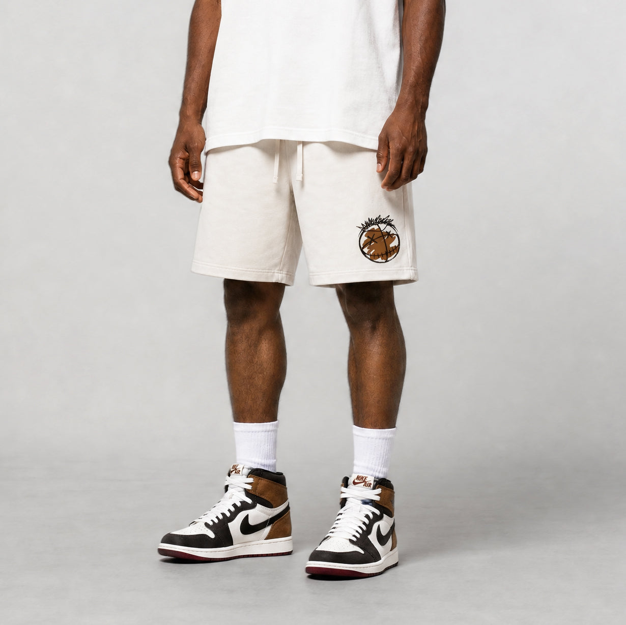 A model wearing off-white Champion shorts with a brown and gold logo patch on the left leg, styled with a white t-shirt, white socks, and brown and white sneakers, against a light gray studio background.