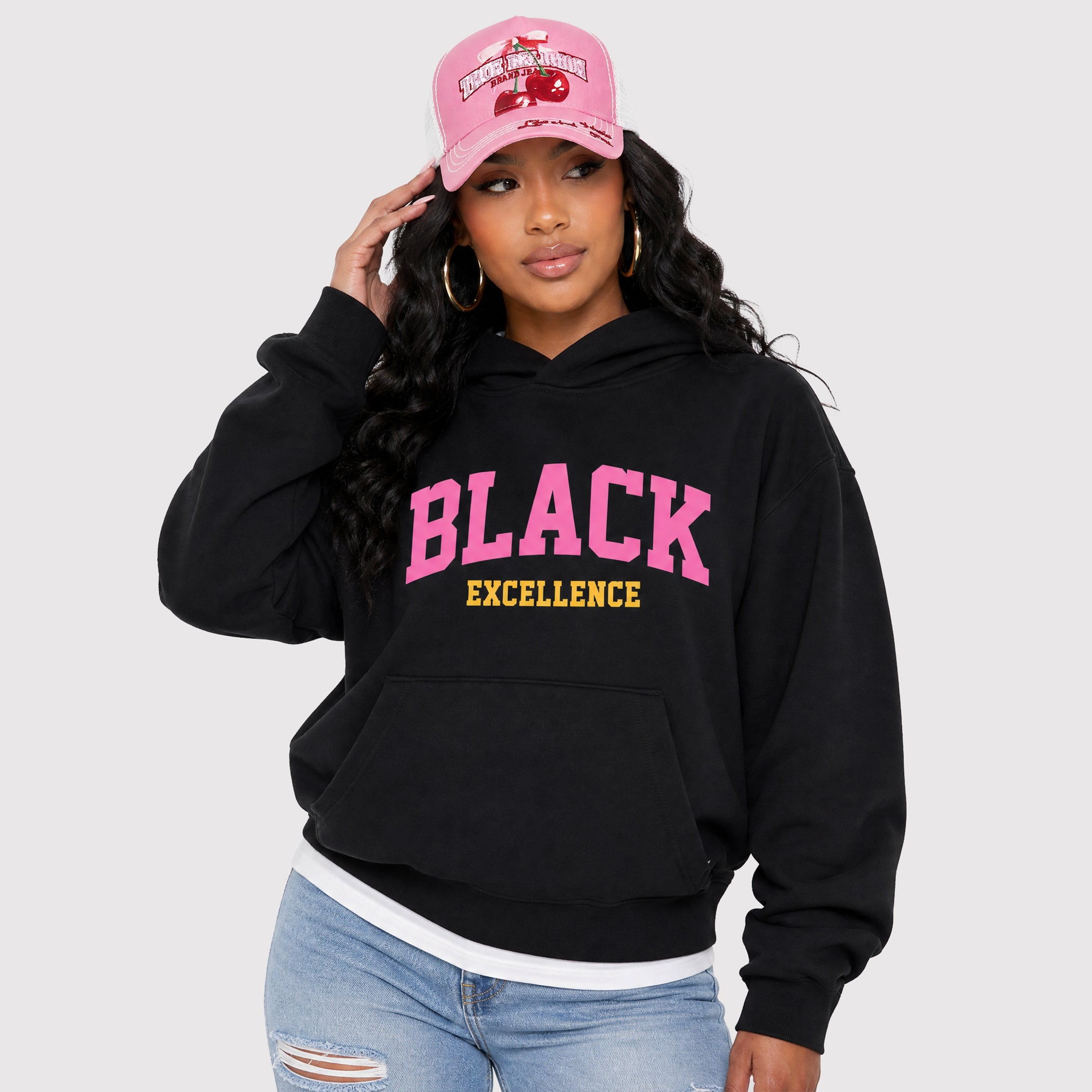 A woman wearing a black pullover hoodie with 'BLACK EXCELLENCE' printed in pink and yellow on the front, paired with a pink baseball cap and light blue jeans, against a light gray studio background.