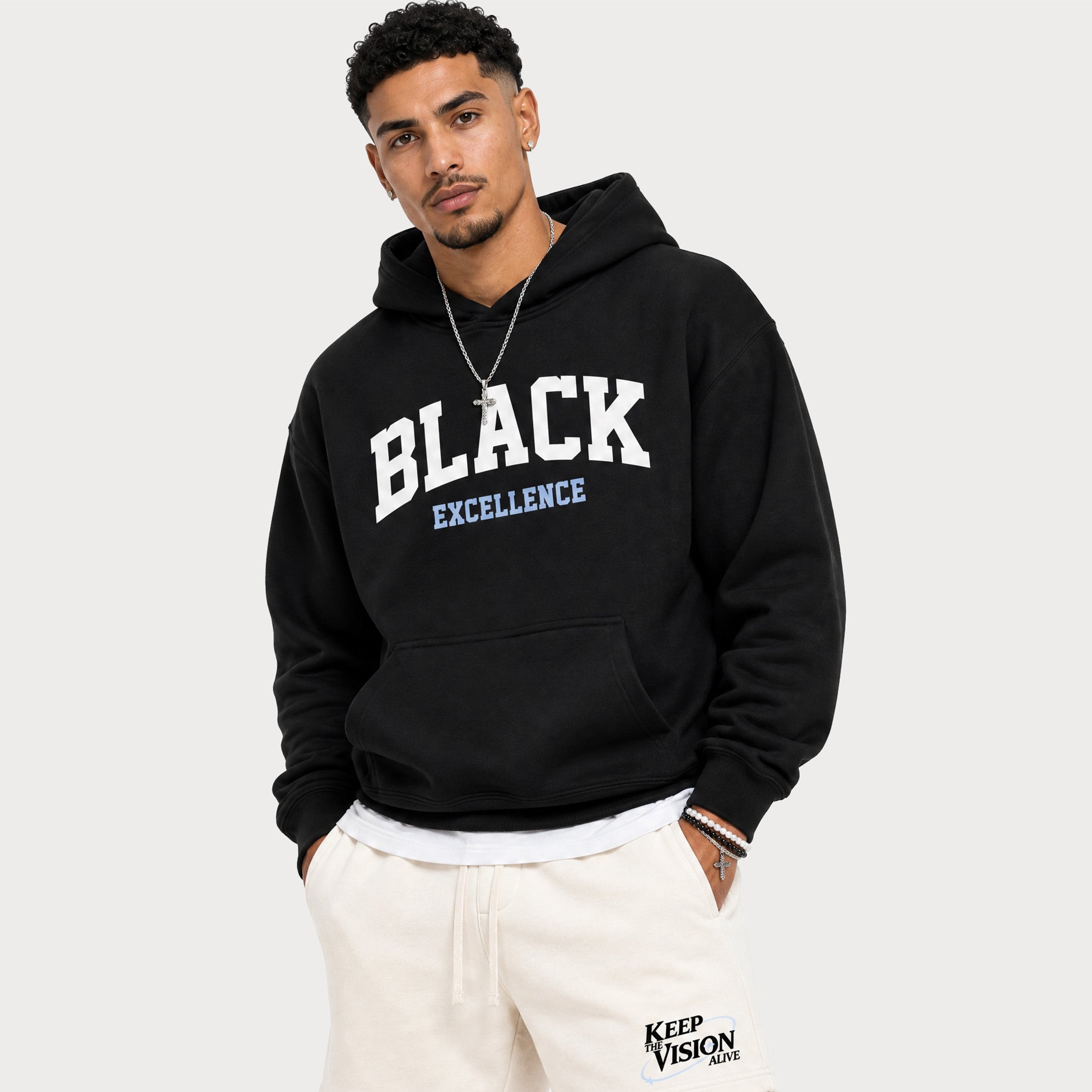 A man wearing a black pullover hoodie with 'BLACK EXCELLENCE' printed in white and light blue on the front, paired with off-white sweatpants, against a light gray studio background.