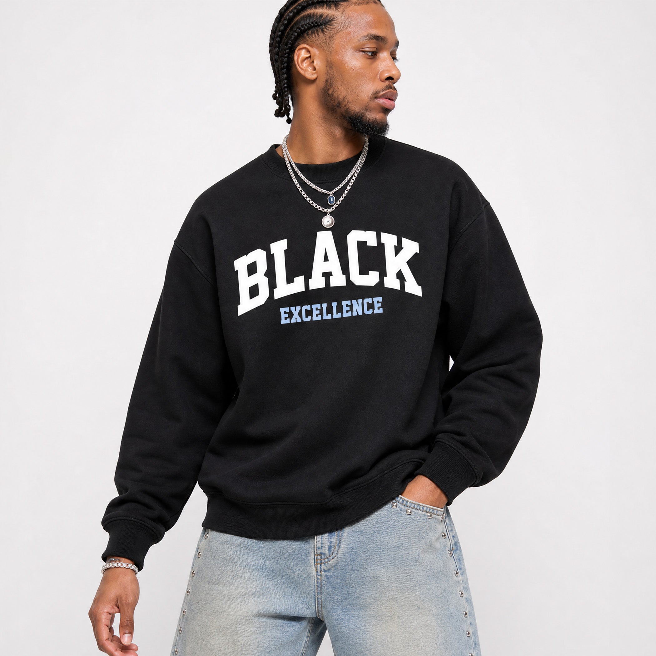 A man wearing a black crewneck sweatshirt with 'BLACK EXCELLENCE' printed in white and light blue on the front, paired with light-wash jeans, against a plain light gray background.