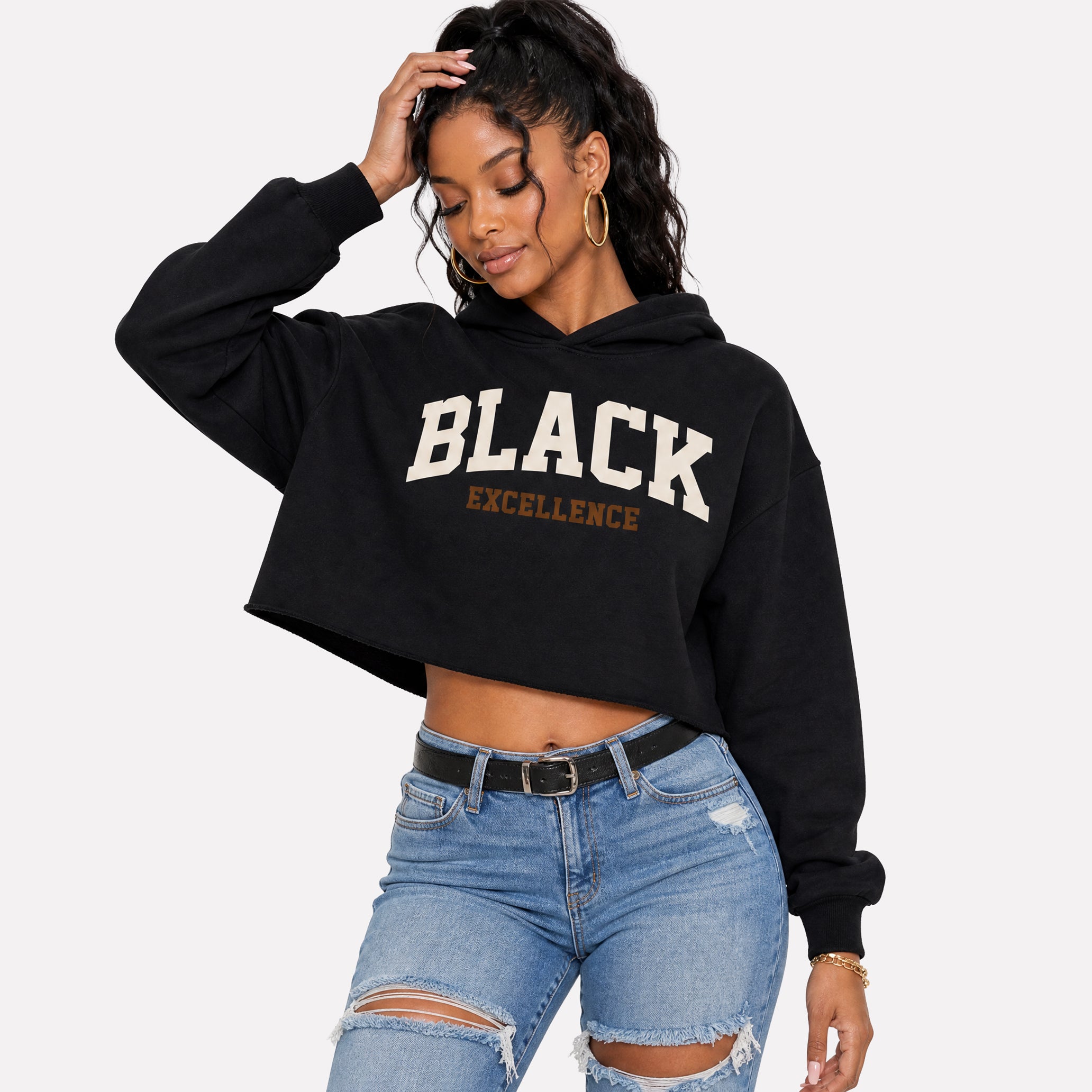 A woman modeling a black cropped hoodie with 'BLACK EXCELLENCE' printed in white and gold on the front, paired with distressed blue jeans, against a light gray background.