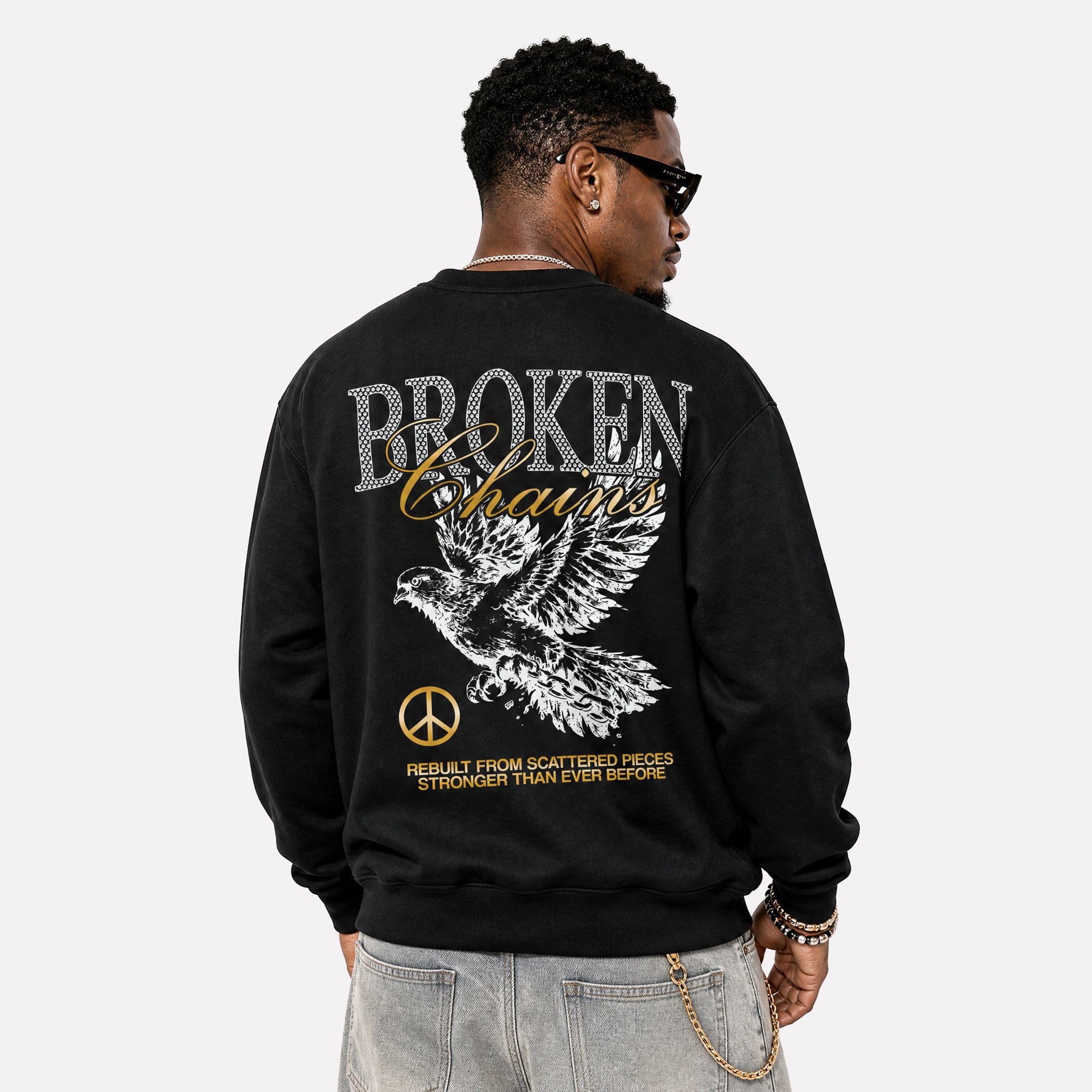 A man wearing a black crewneck sweatshirt from the back, featuring a large graphic print with the words 'BROKEN Chains', a white eagle, and gold and silver text on a light gray background.