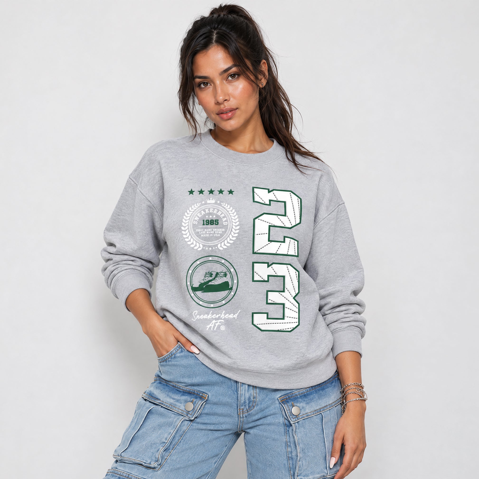 A woman modeling a light gray crewneck sweatshirt with a green and white graphic print featuring the number 23 and circular emblems, paired with light blue jeans against a light gray studio background.
