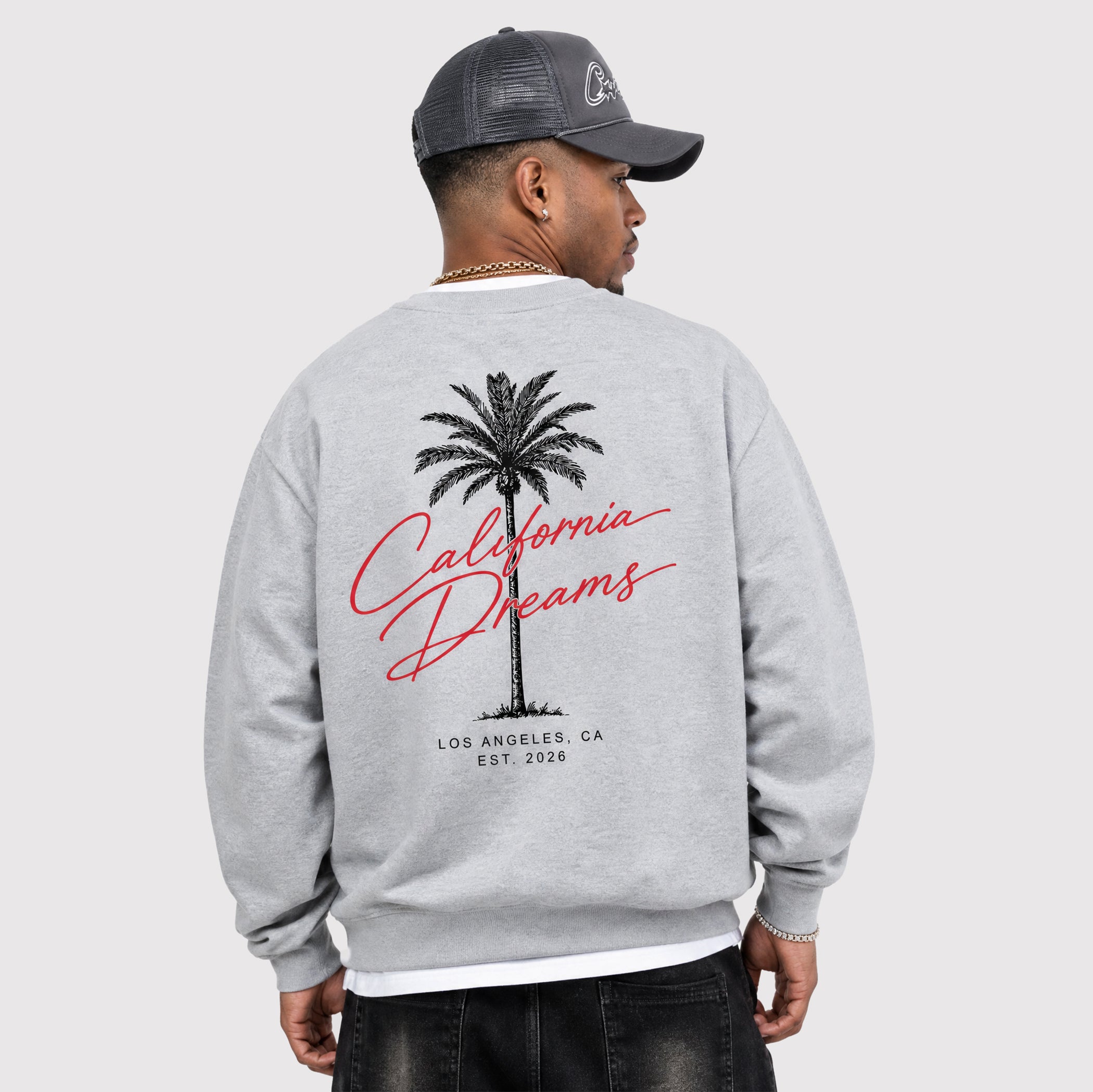 A man shown from the back wearing a light gray crewneck sweatshirt with a graphic print of a palm tree and the words 'California Dreams' in red and black. He is also wearing a dark gray trucker hat and dark jeans, set against a plain light gray background.