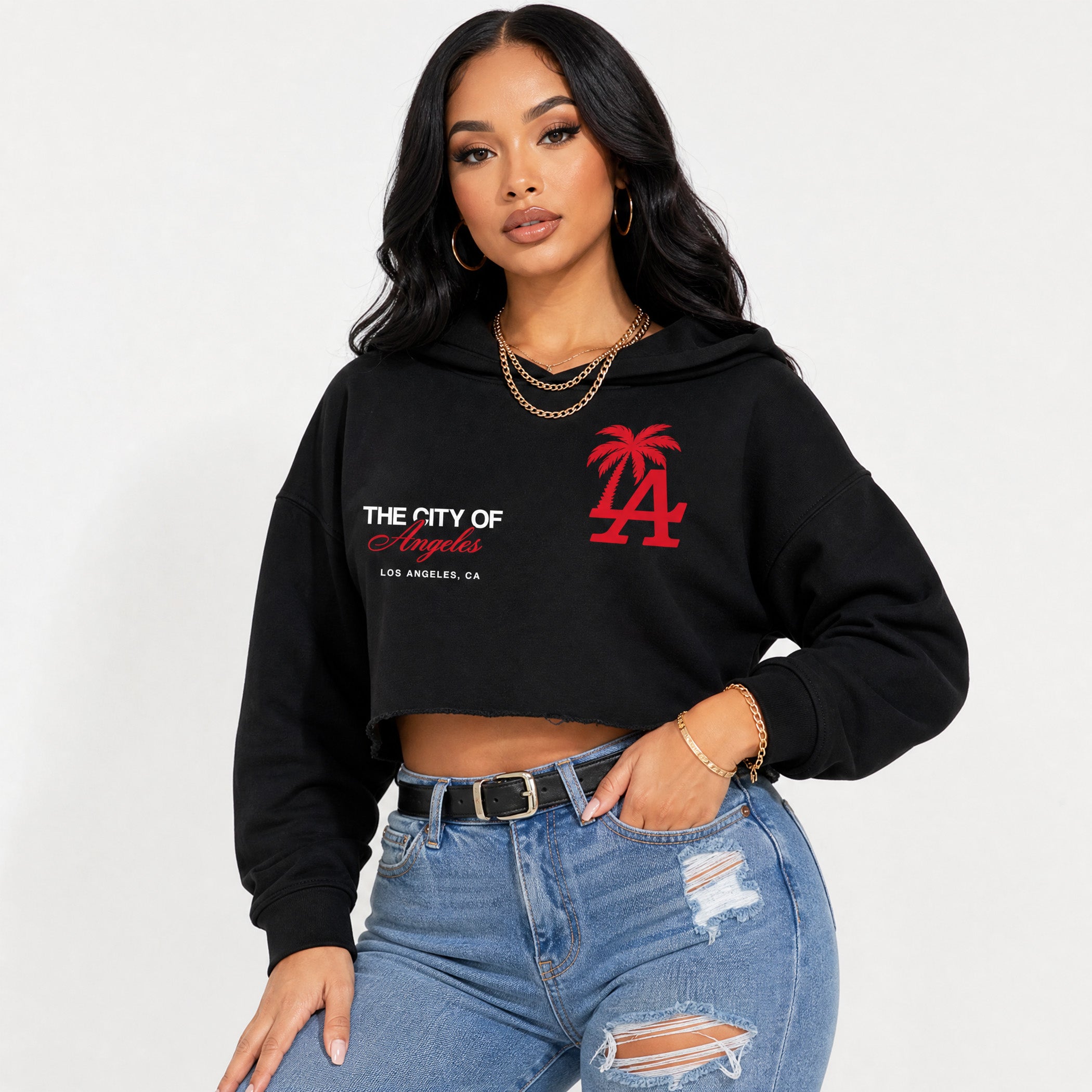 A woman modeling a black cropped hoodie with red and white graphic text and logo on the front, paired with distressed blue jeans, against a plain light gray studio background.
