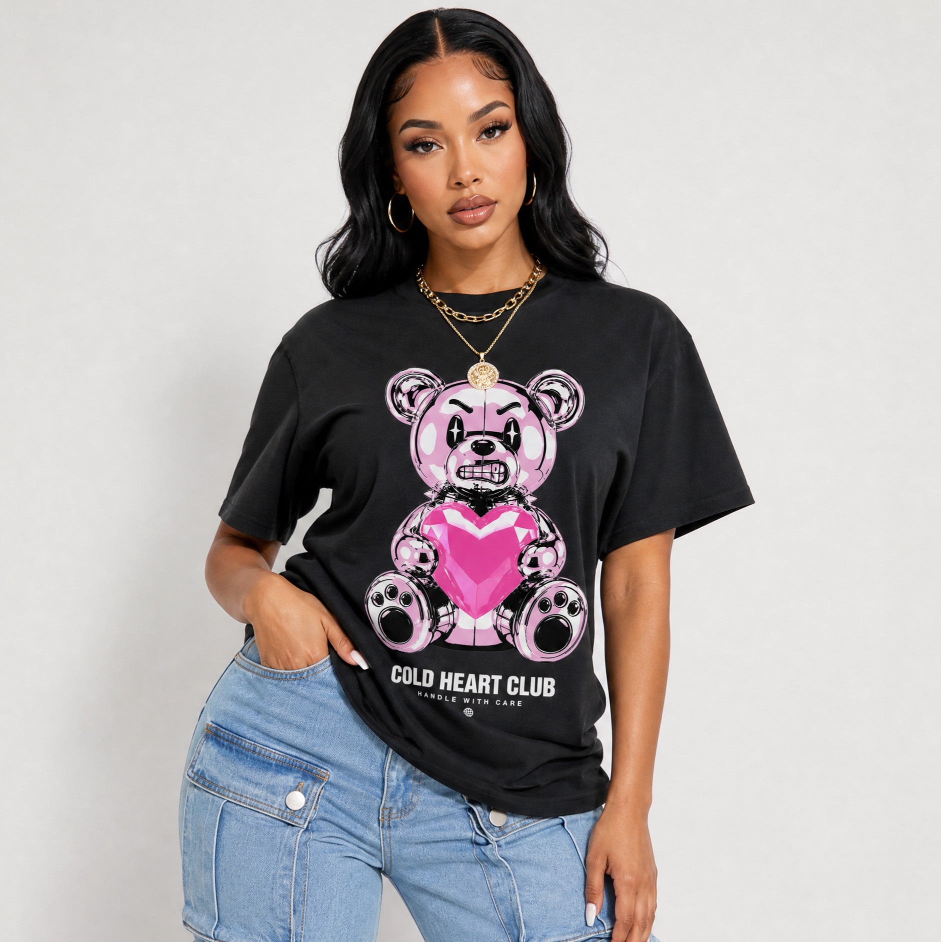 A woman modeling a black graphic t-shirt featuring a pink teddy bear with a heart and the text 'GOLD HEART CLUB'. She is wearing light blue denim jeans and gold jewelry, posing against a light gray studio background.