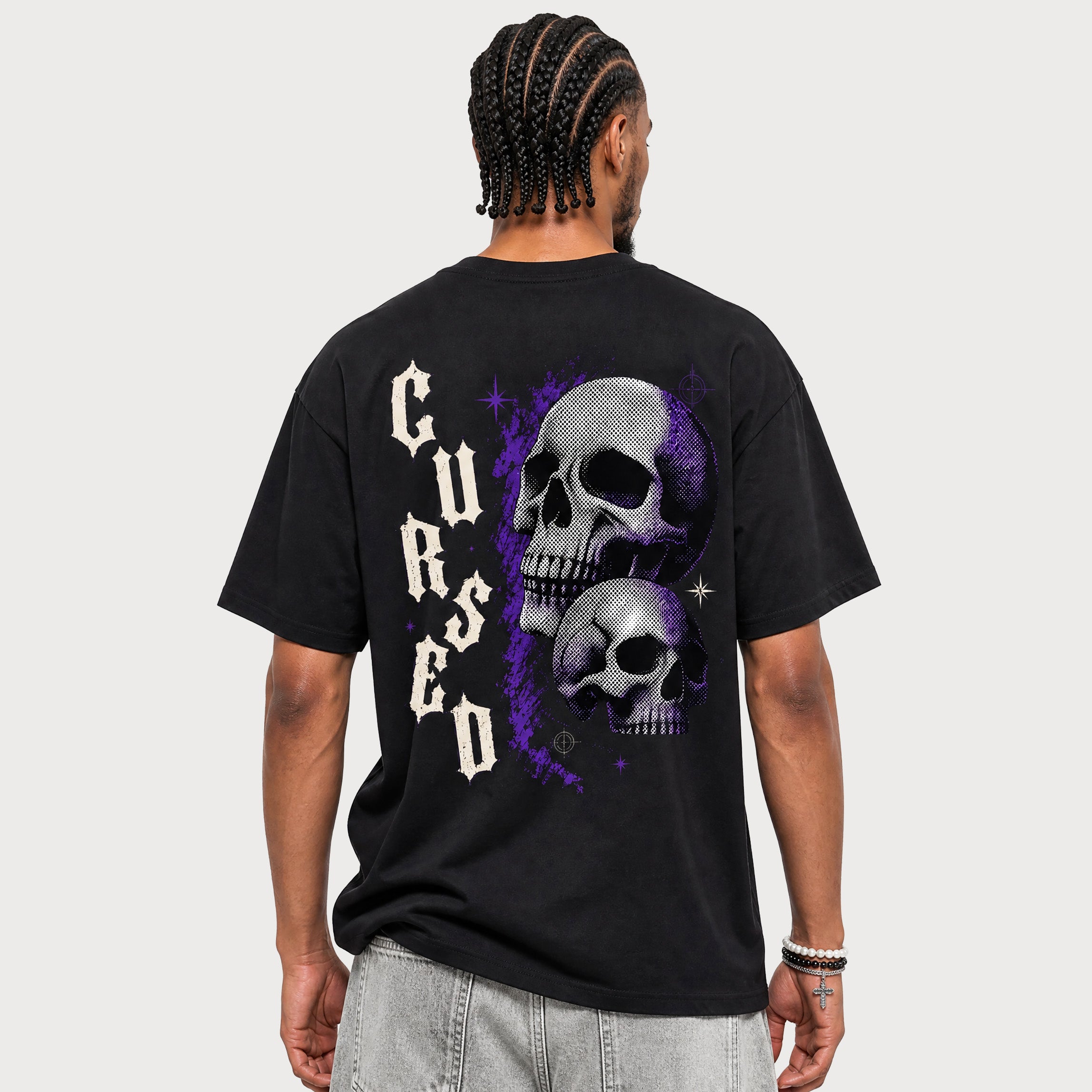 A man viewed from the back wearing a black t-shirt with a large graphic print featuring two skulls and the word 'Cursed' in a gothic font, paired with light gray jeans against a light gray background.