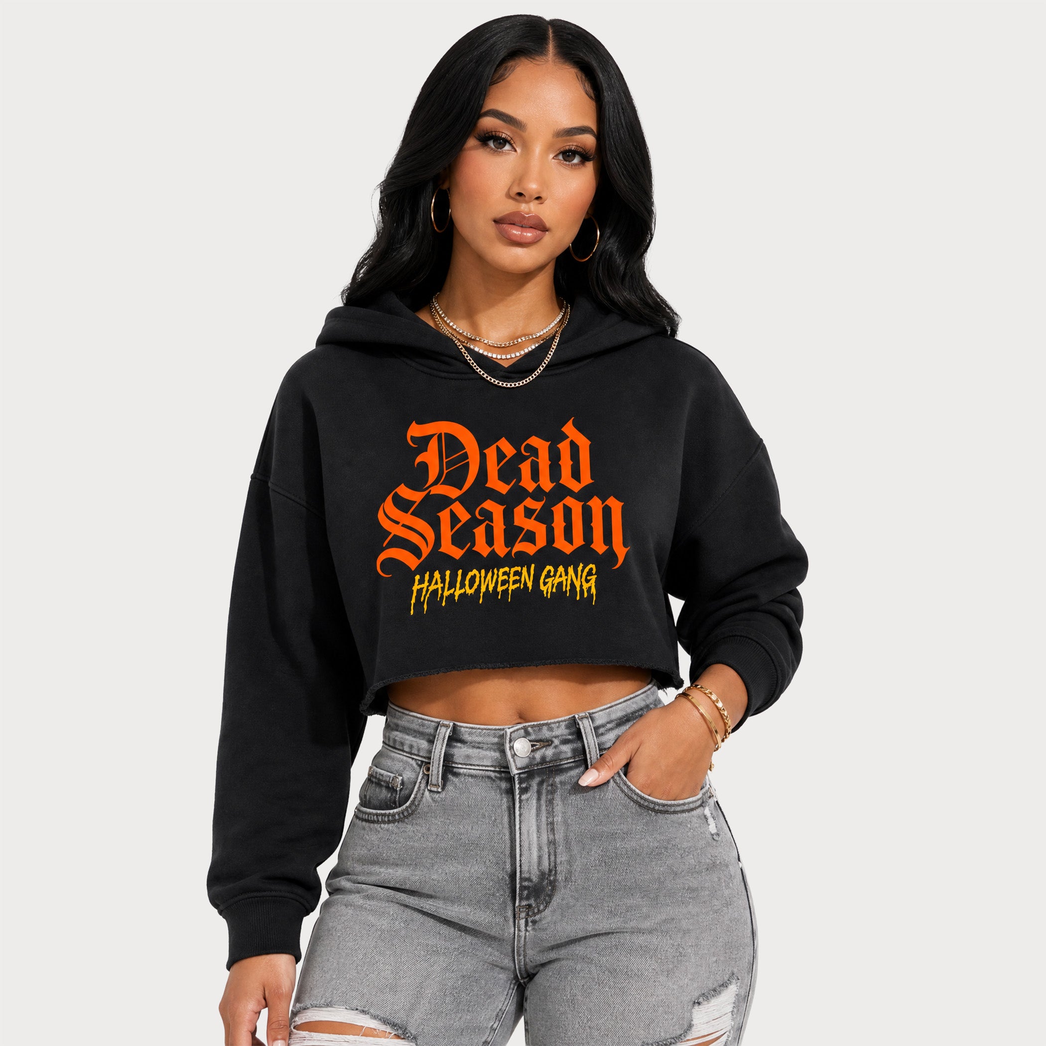 A woman modeling a black cropped hoodie with 'Dead Season Halloween Gang' printed in orange and yellow gothic-style text on the front, paired with light gray distressed jeans against a light gray studio background.