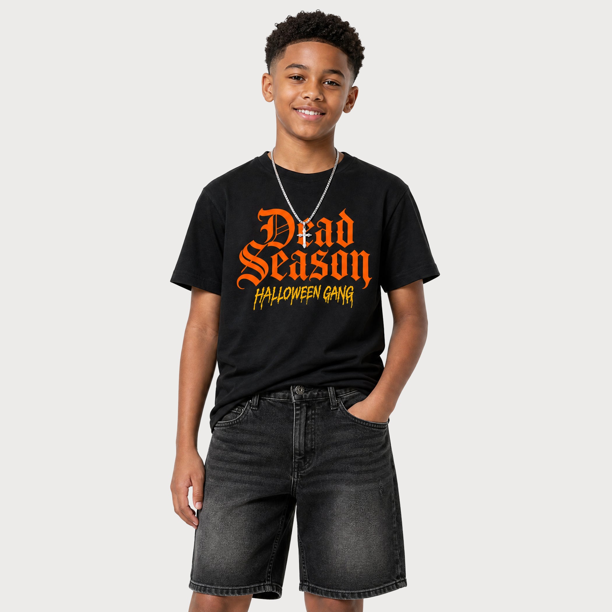 A young man wearing a black graphic t-shirt with 'Dead Season Halloween Gang' printed in orange and yellow gothic font, paired with dark gray denim shorts, against a solid light gray background.