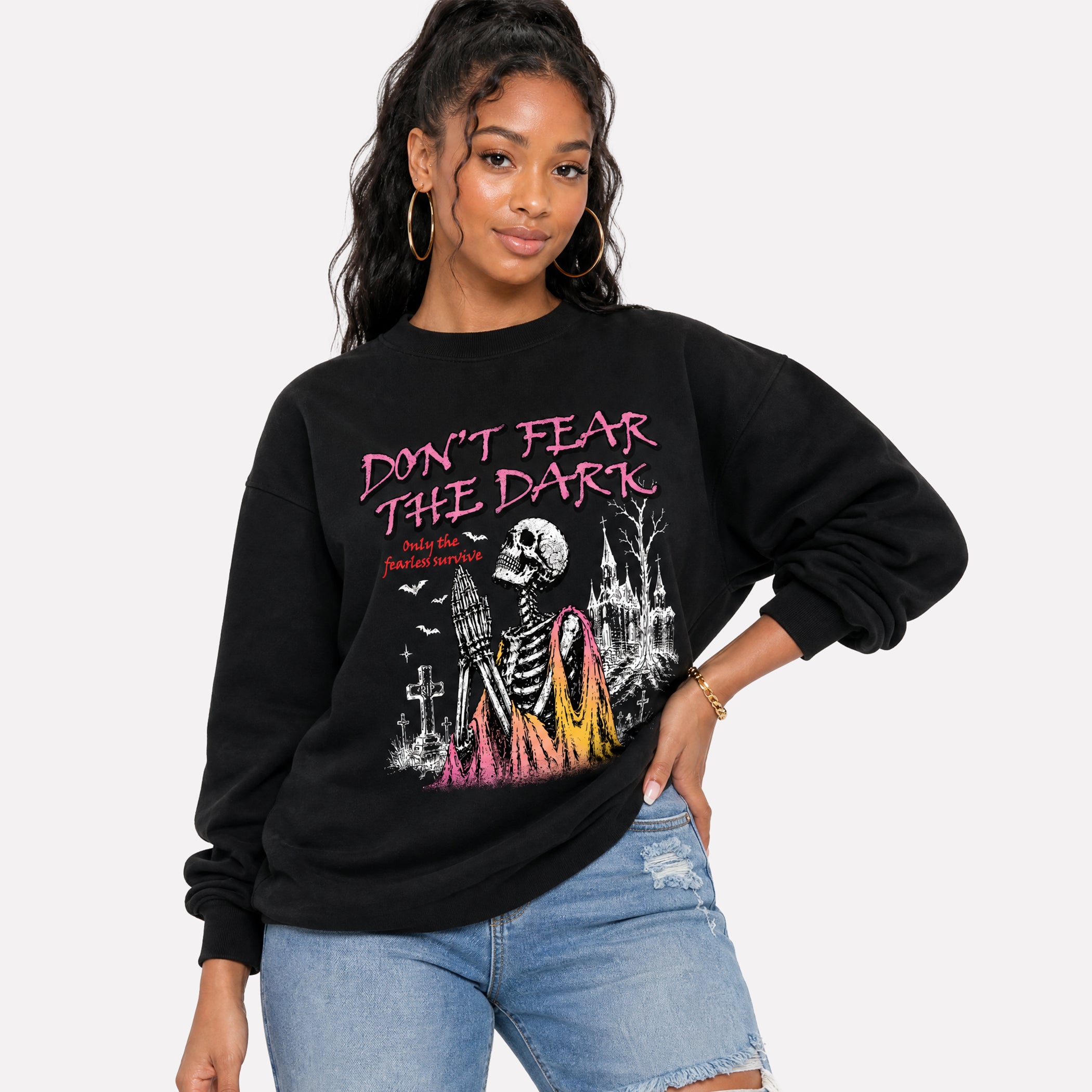 A woman modeling a black crewneck sweatshirt with a colorful gothic graphic featuring a skeleton, a ribbon, and the text 'DON'T FEAR THE DARK' in pink, paired with light blue denim shorts against a light gray background.