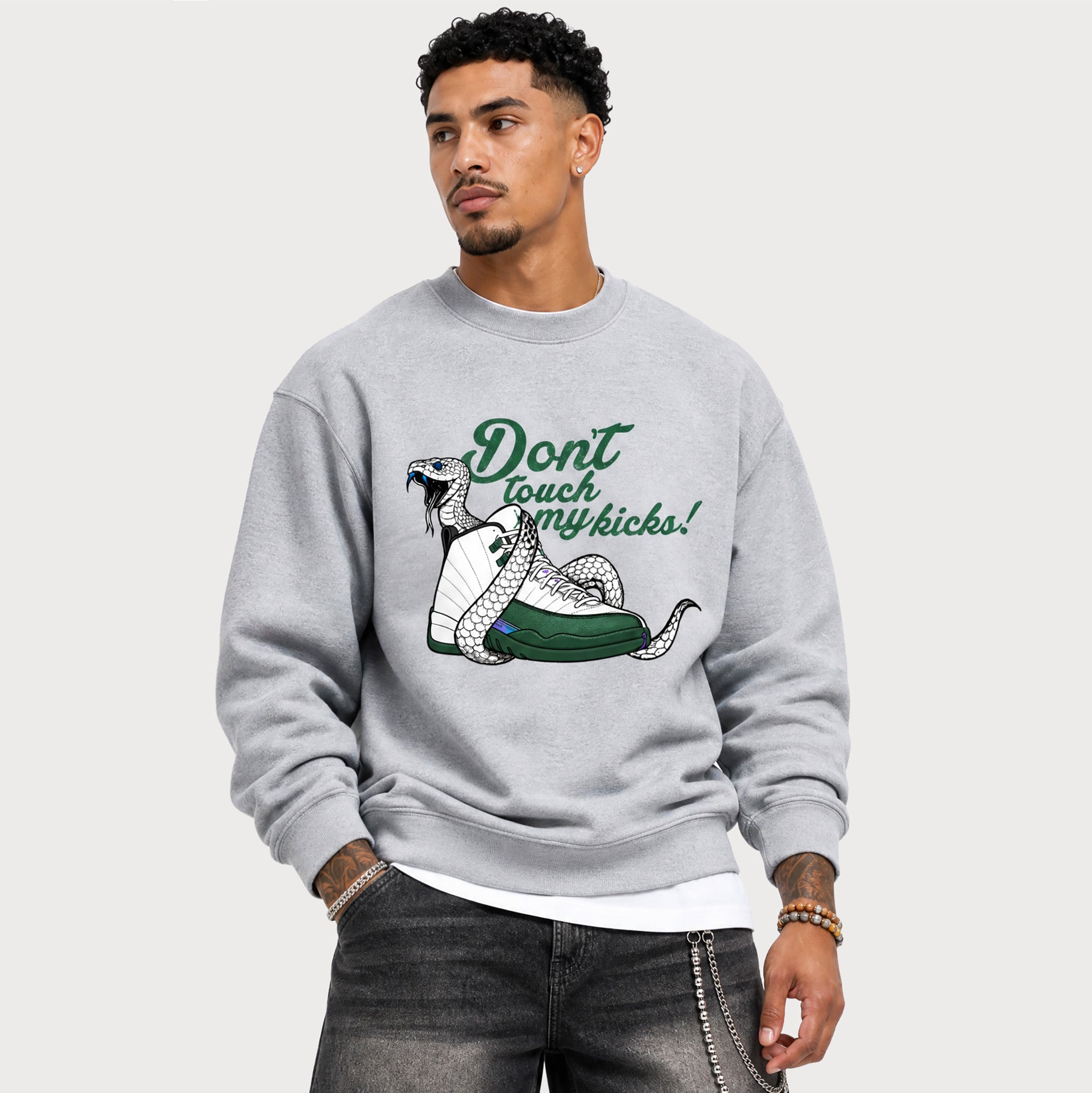 A man wearing a light gray crewneck sweatshirt with a graphic of a coiled snake and the text 'Don't touch my kicks!' in green and black, paired with dark gray jeans against a light gray studio background.