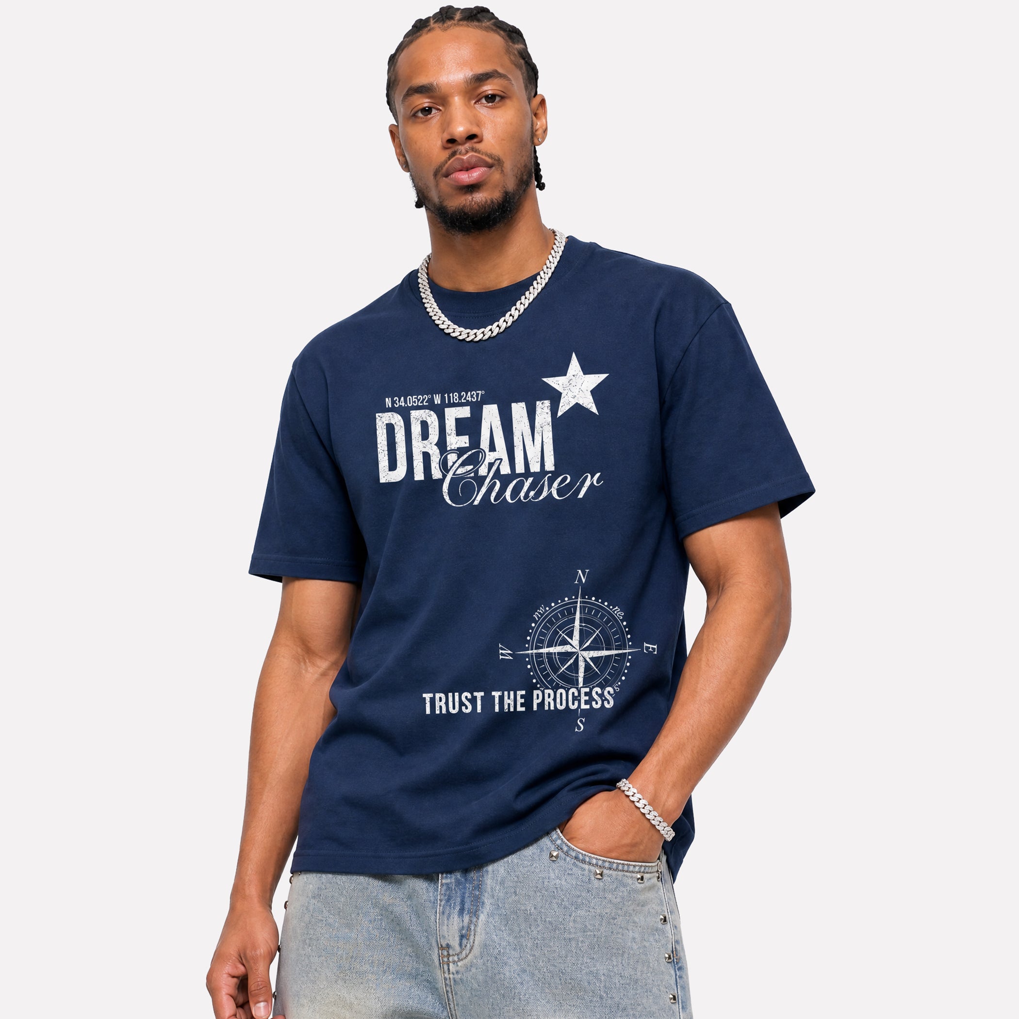 A man wearing a navy blue graphic t-shirt with 'Dream Chaser' and a compass design printed on the front, paired with light-wash jeans against a light gray background.