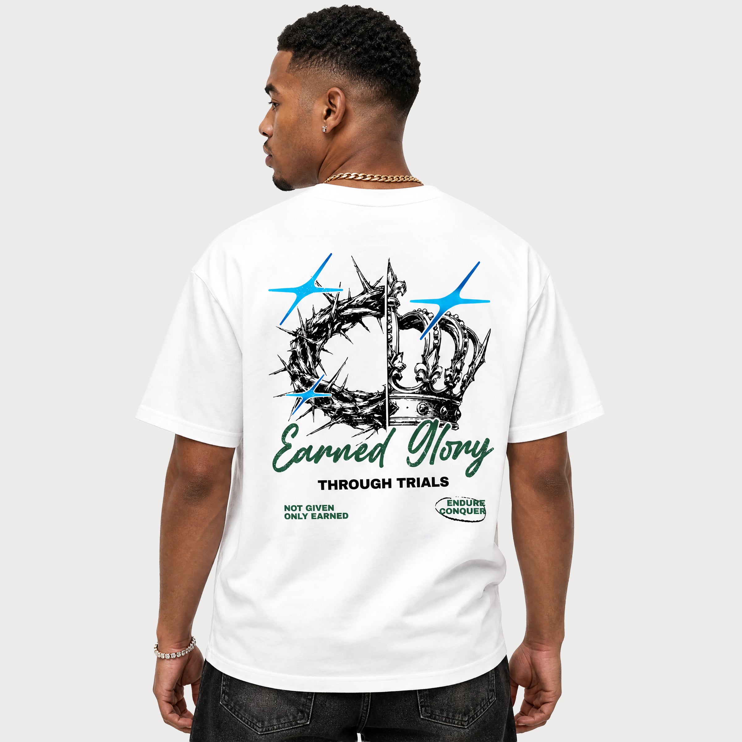 A man wearing a white graphic t-shirt, viewed from the back. The shirt features a large graphic print with a crown of thorns, two blue airplanes, and the text 'Earned Glory THROUGH TRIALS' in black and green, along with 'NOT GIVING ONLY EARNED' and a '4 HOURS REMOVAL' label.
