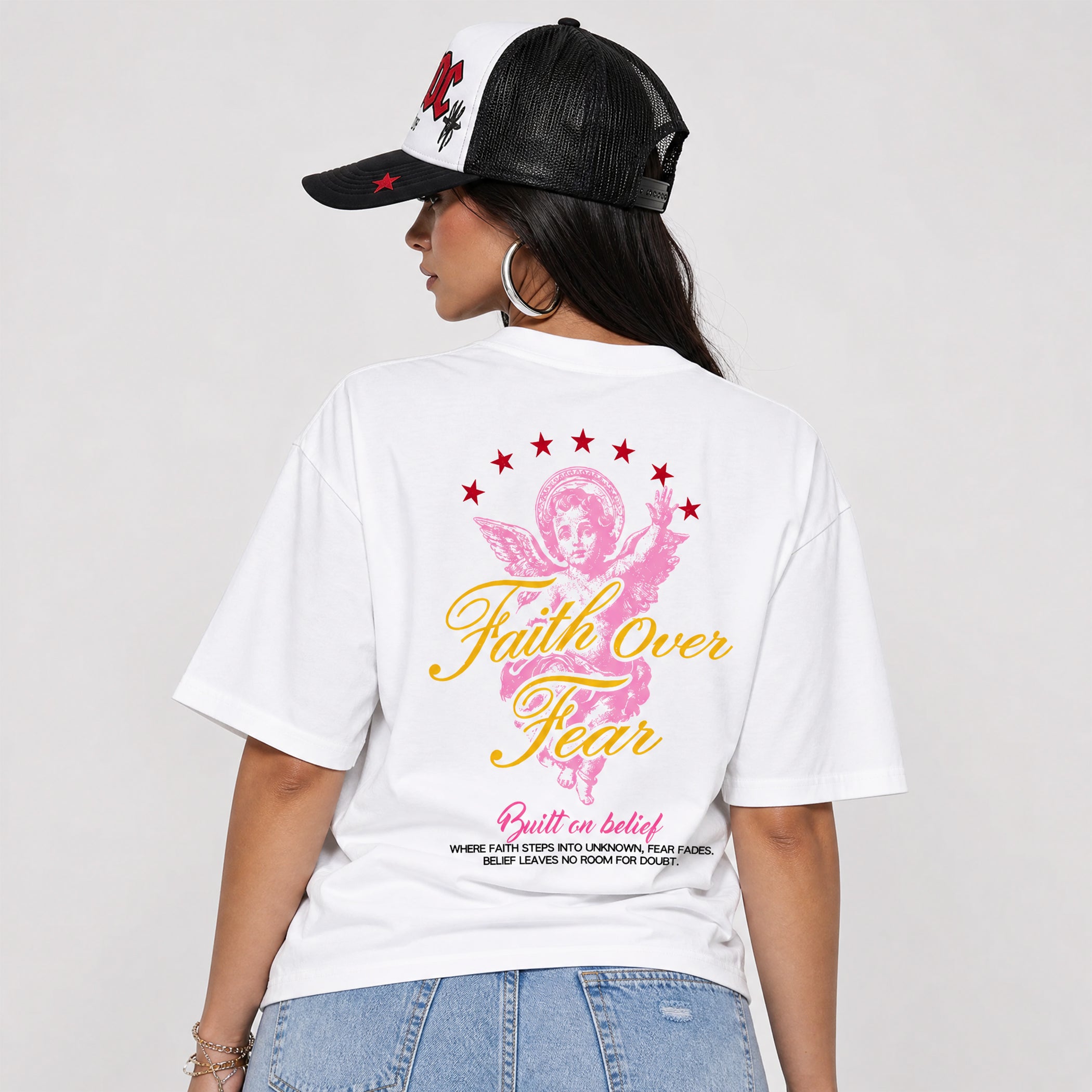 A woman shown from the back wearing a white graphic T-shirt and a black-and-white trucker hat. The shirt features a pink angel graphic with red stars and yellow text reading 'Faith Over Fear' on a light gray studio background.