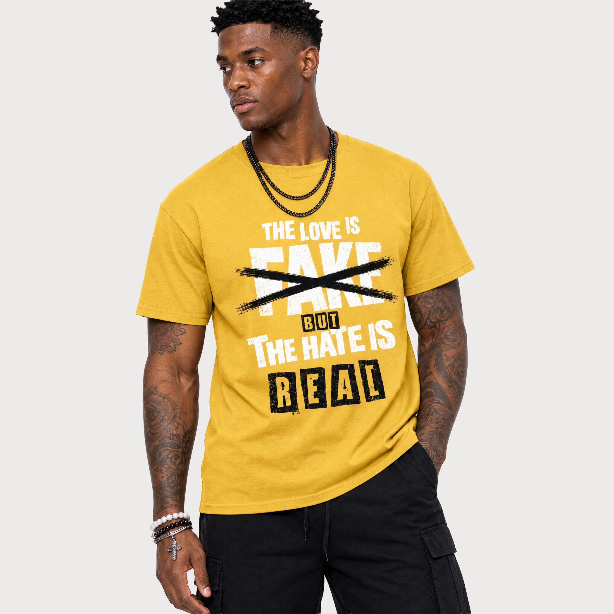 A man wearing a yellow graphic t-shirt with the text 'THE LOVE IS FAKE BUT THE HATE IS REAL' in white and black, paired with black cargo pants, against a light gray background.
