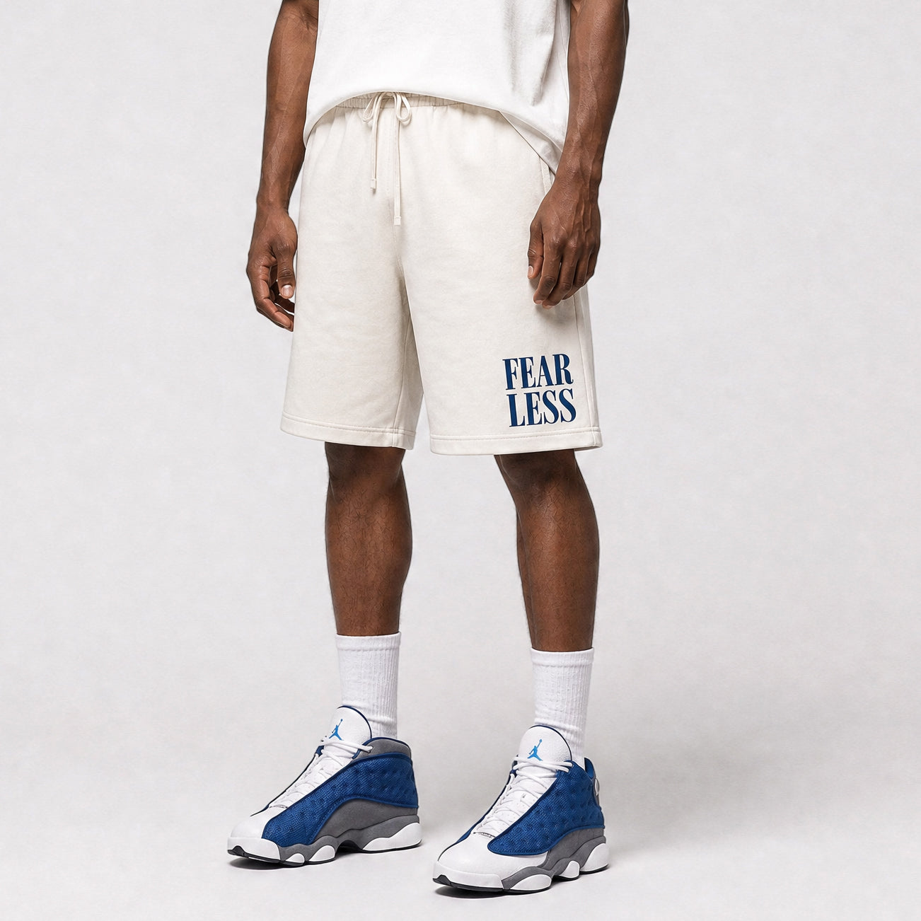 A model wearing off-white sweat shorts with a blue 'FEAR LESS' graphic on the left leg, paired with white socks and blue and white sneakers, against a light gray studio background.