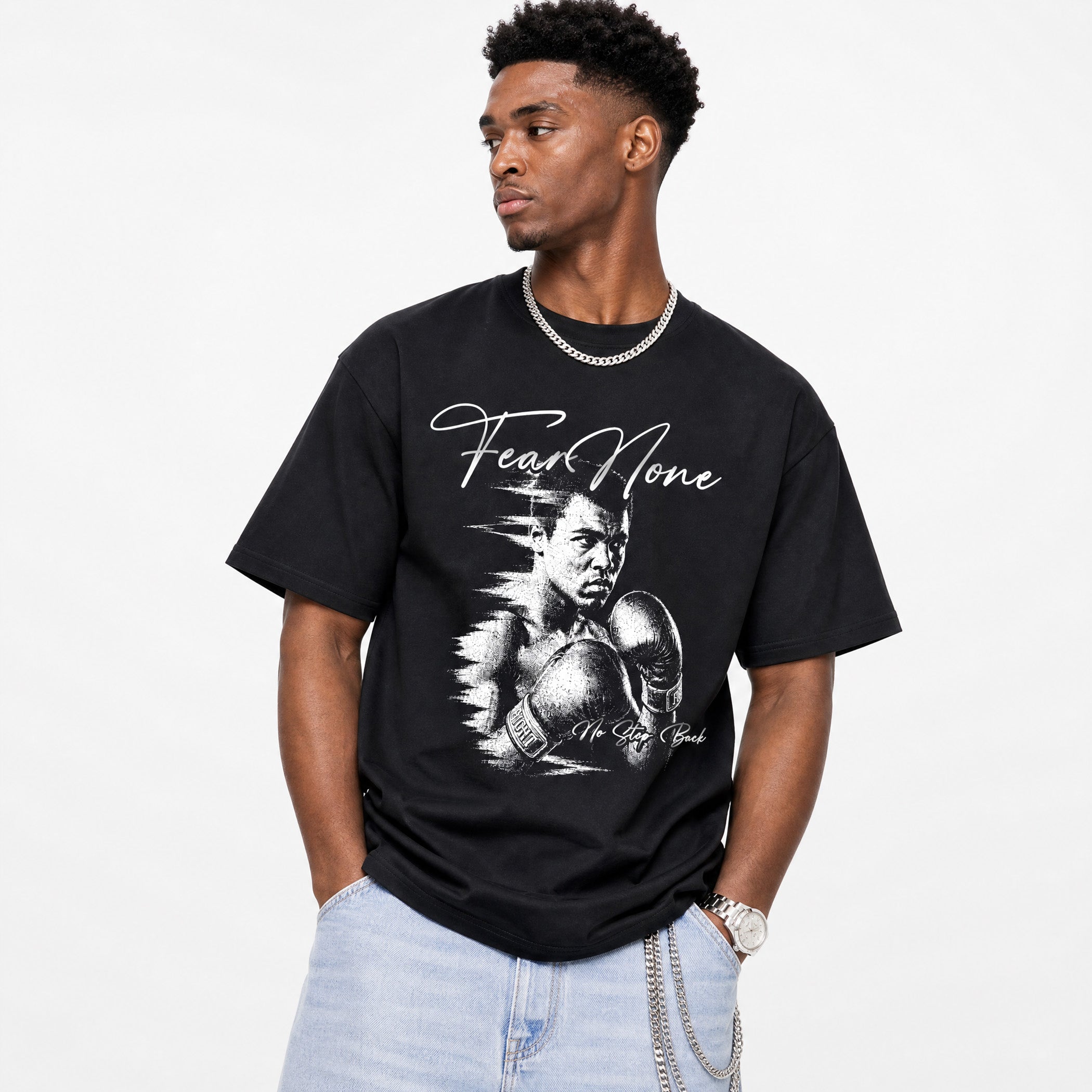 A man wearing a black graphic t-shirt featuring a white sketch of Muhammad Ali and the text 'Fear None' on the front, paired with light blue jeans and a silver chain necklace, set against a plain white background.