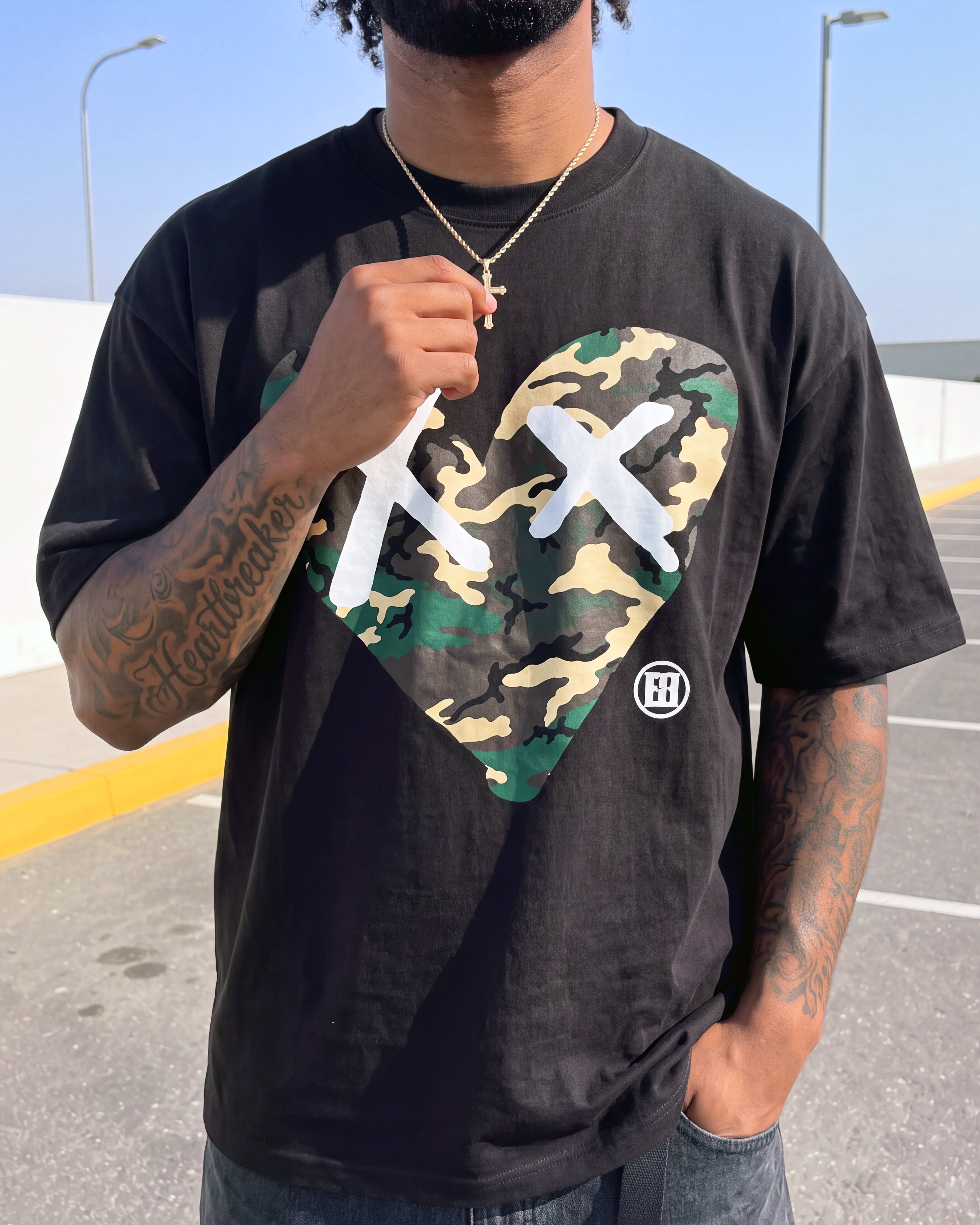 A man wearing a black graphic t-shirt featuring a heart-shaped camouflage print with white X's and a small circular logo, photographed outdoors in a parking lot.