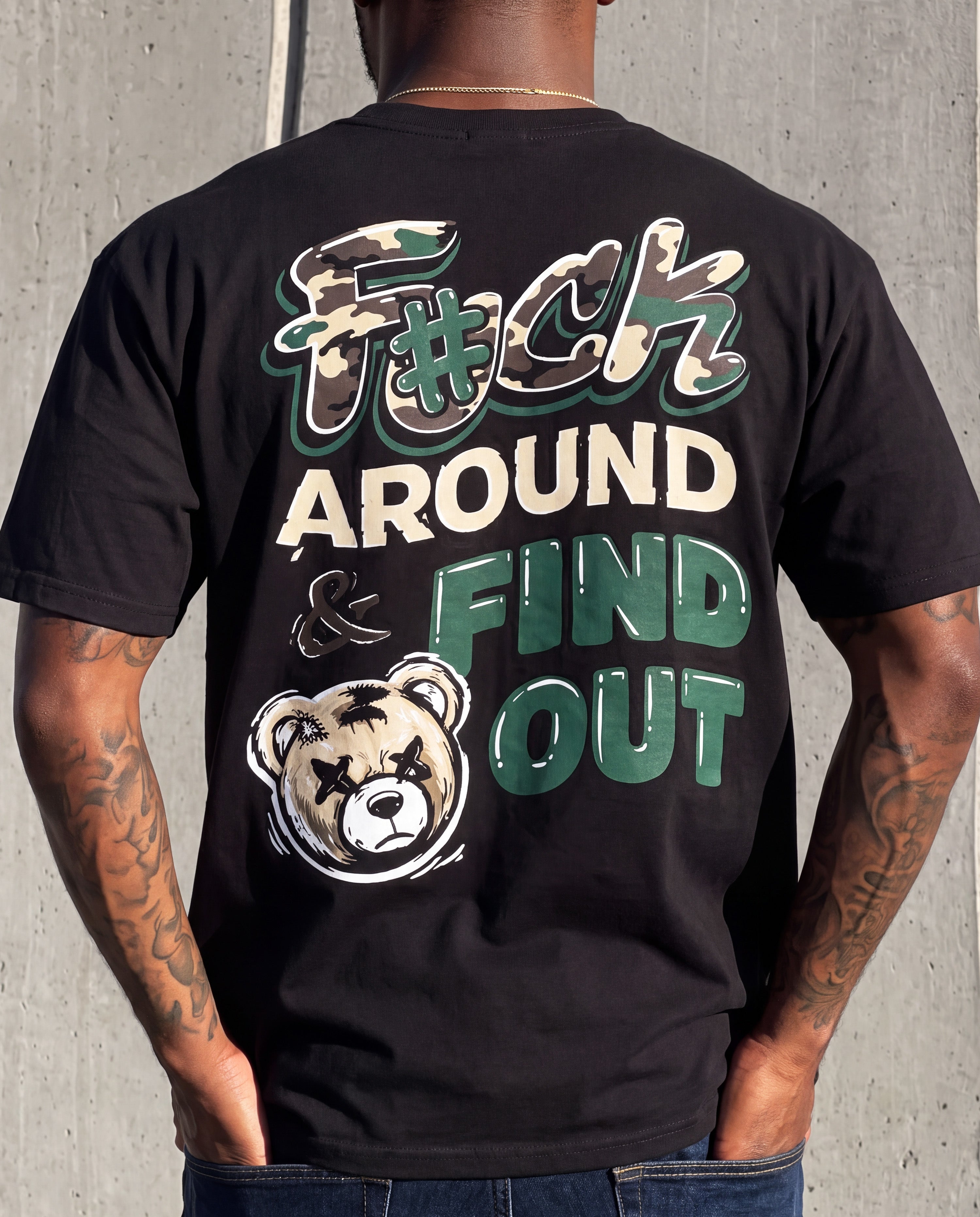 A man wearing a black graphic t-shirt, viewed from the back. The shirt features a bold design with the text 'F*#CK AROUND & FIND OUT' in green, beige, and white, alongside a cartoon bear graphic.