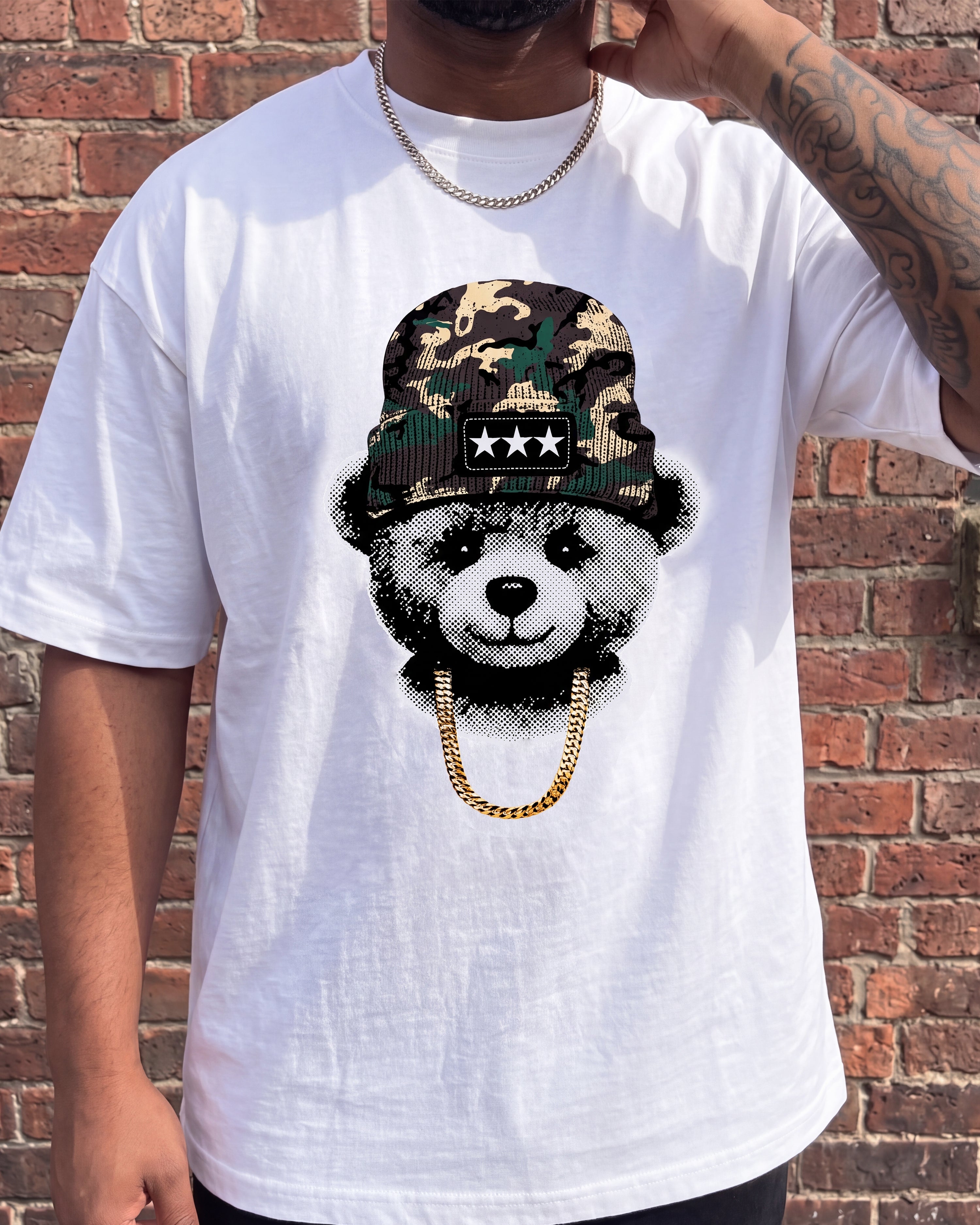 A man wearing a white graphic t-shirt featuring a teddy bear wearing a camouflage bucket hat and a gold chain, posed against a red brick wall.