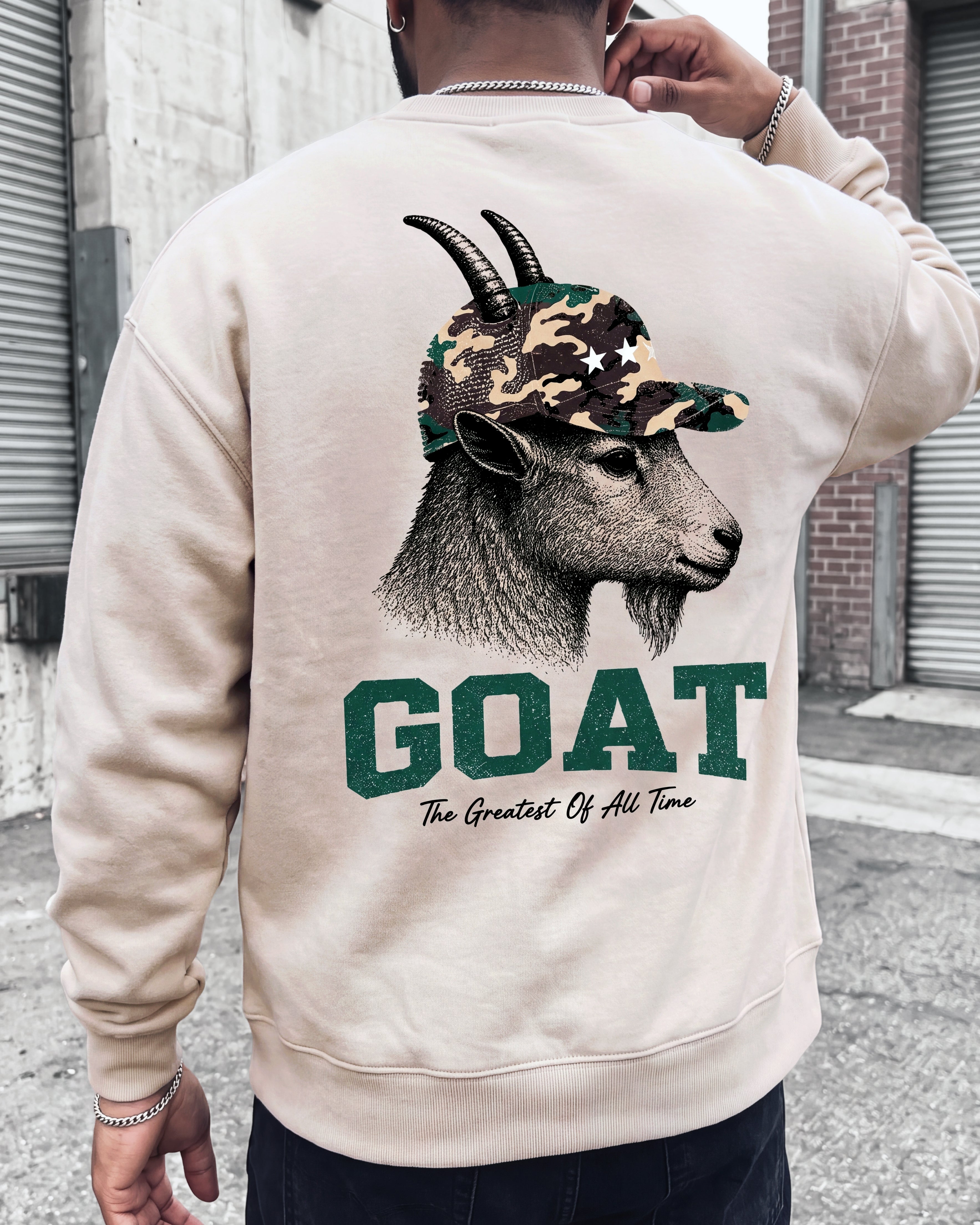 A man wearing a beige crewneck sweatshirt, viewed from the back. The sweatshirt features a large graphic print of a goat wearing a camouflage baseball cap, with the word 'GOAT' in bold green letters and the phrase 'The Greatest Of All Time' underneath.