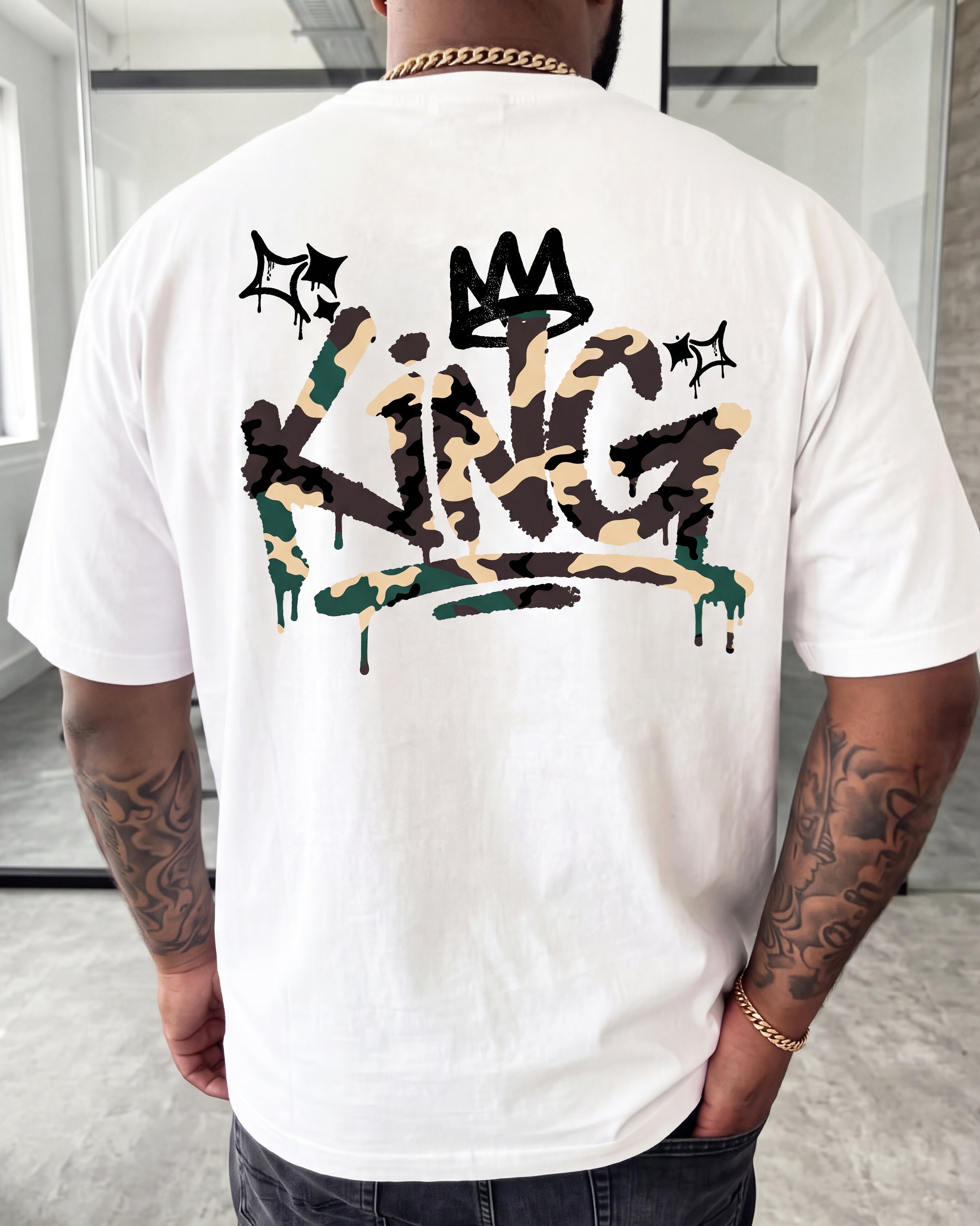 A man wearing a white t-shirt from the back, featuring a large graphic print of the word 'KING' in a camouflage spray-paint style with a black crown above it.