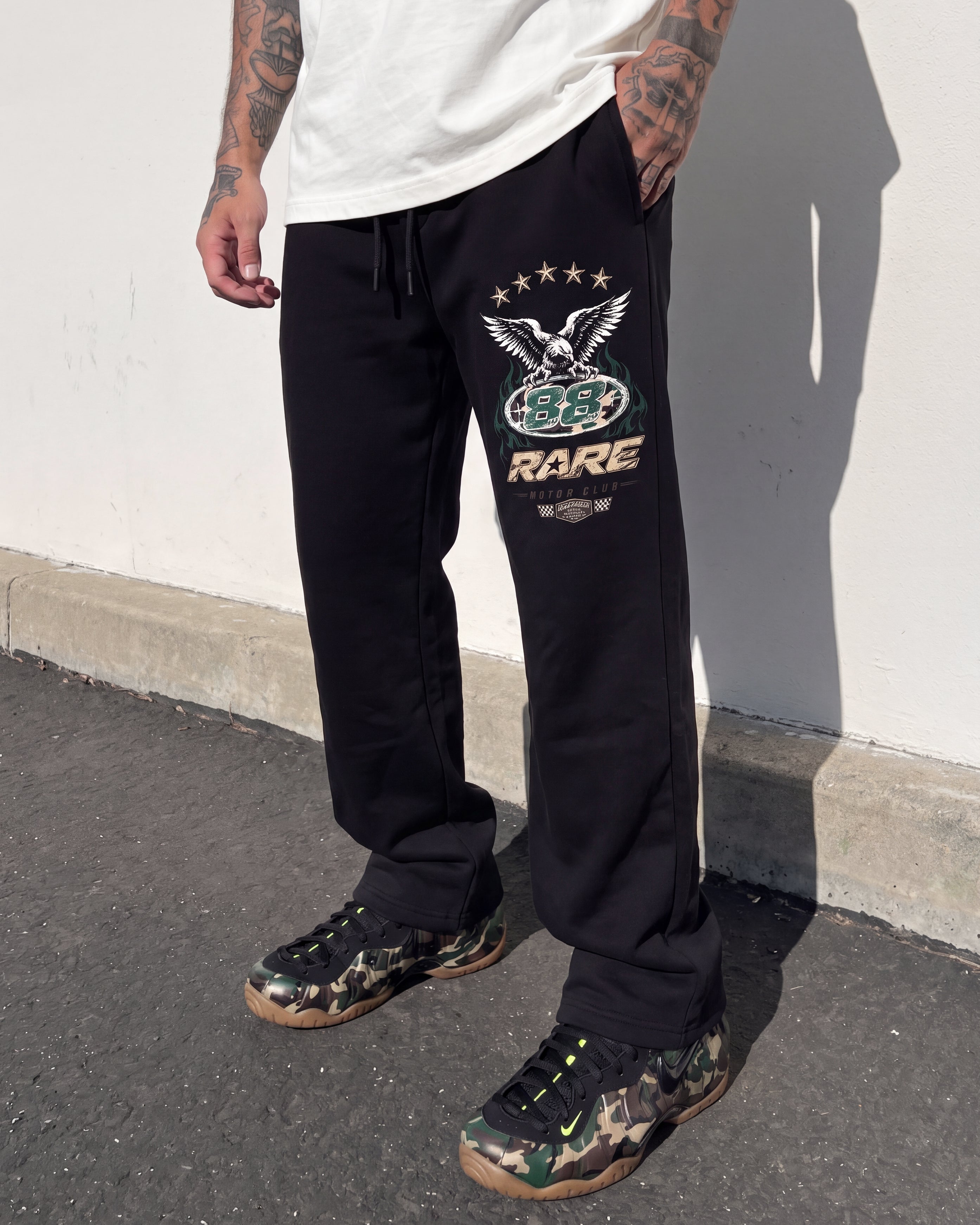 A person wearing black sweatpants with a large graphic print on the left thigh, paired with camouflage sneakers and a white t-shirt, standing outdoors against a white wall.