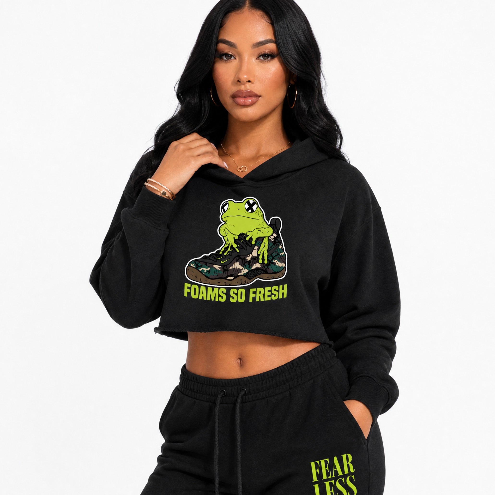 A woman modeling a black cropped hoodie with a green frog graphic sitting on a sneaker and the text 'FOAMS SO FRESH' in neon green, paired with black sweatpants featuring 'FEAR LESS' in yellow-green text.