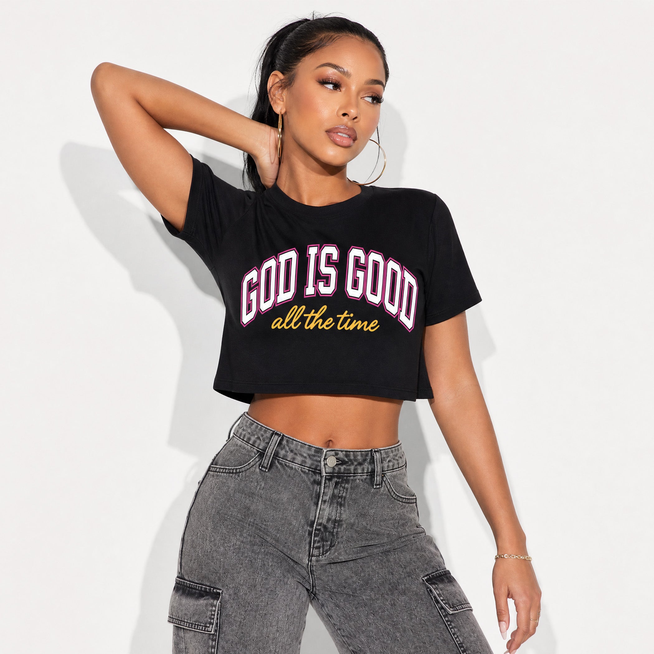 A woman modeling a black cropped t-shirt with 'GOD IS GOOD all the time' printed on the front in white and yellow, paired with gray cargo jeans against a plain white background.