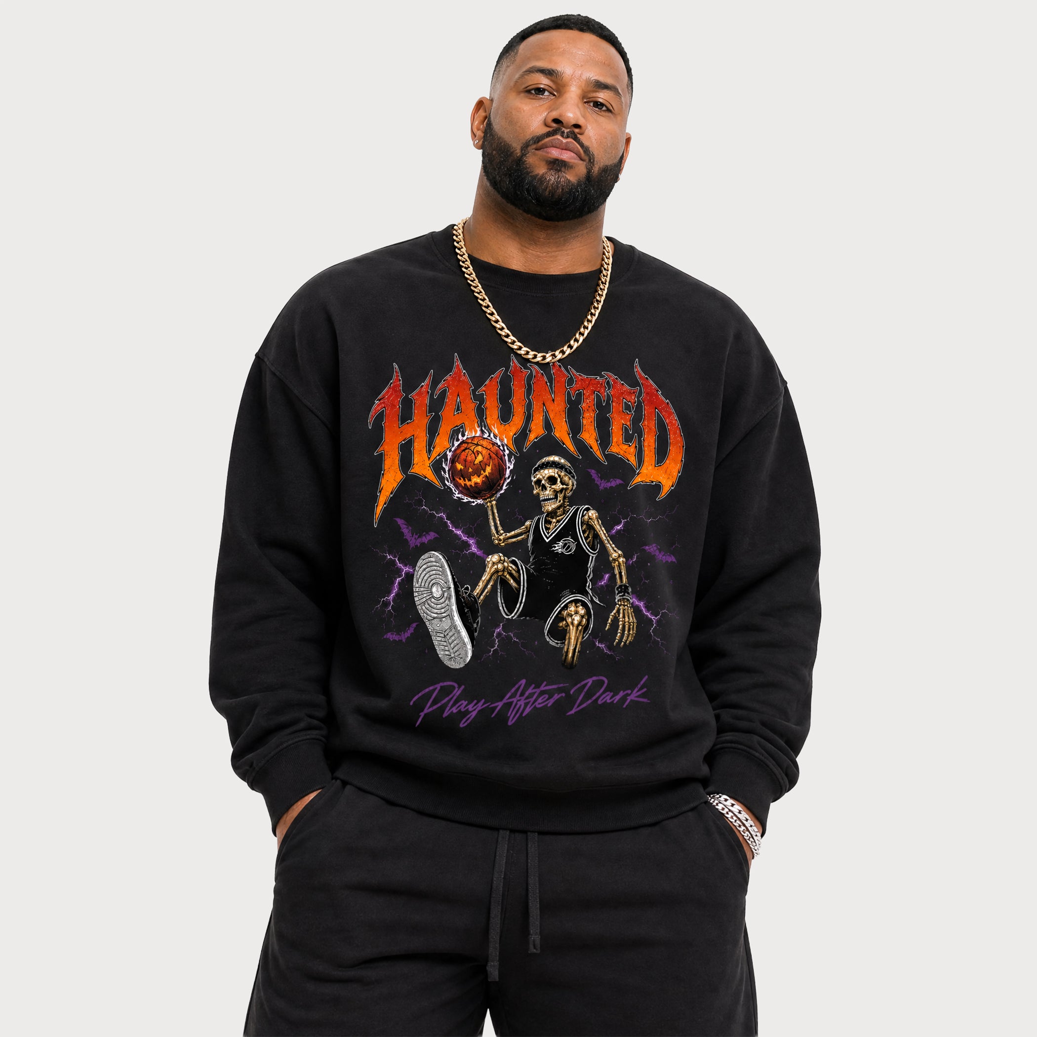 A man wearing a black crewneck sweatshirt with a graphic print of a skeleton, a basketball, a shoe, and the text 'HAUNTED' and 'Play After Dark' in orange, purple, and white. He is also wearing black sweatpants and a gold chain necklace, photographed against a solid light gray background.