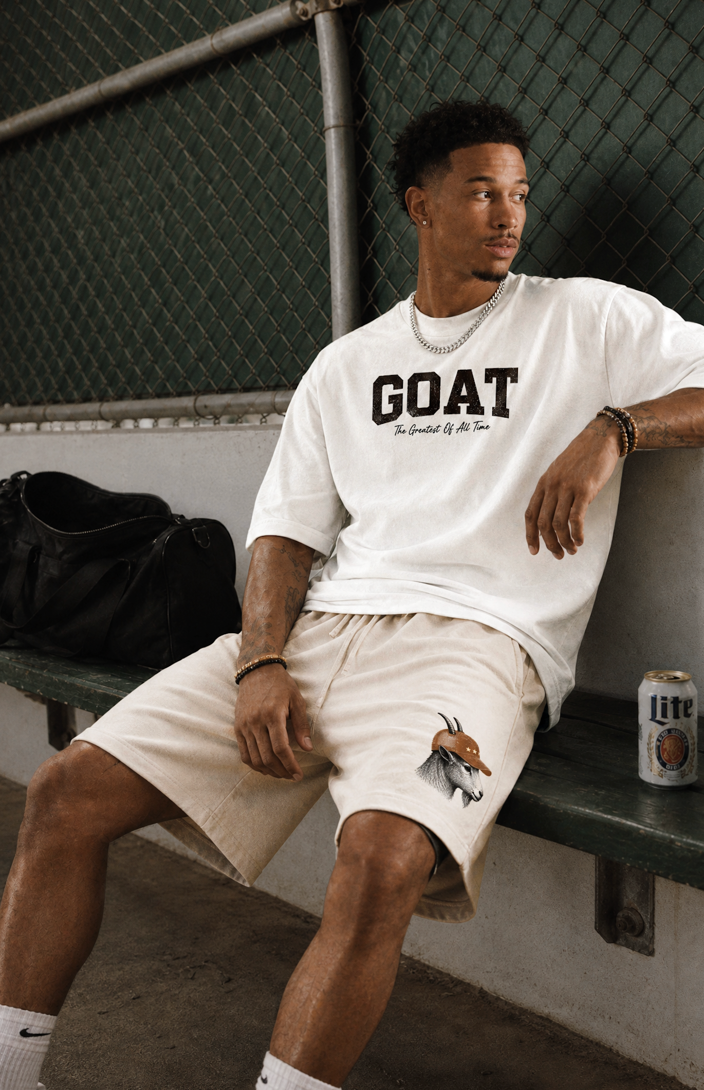 A man sitting on a bench in a dimly lit indoor setting wearing a white GOAT t-shirt and beige shorts, with a black bag and a beer can nearby.