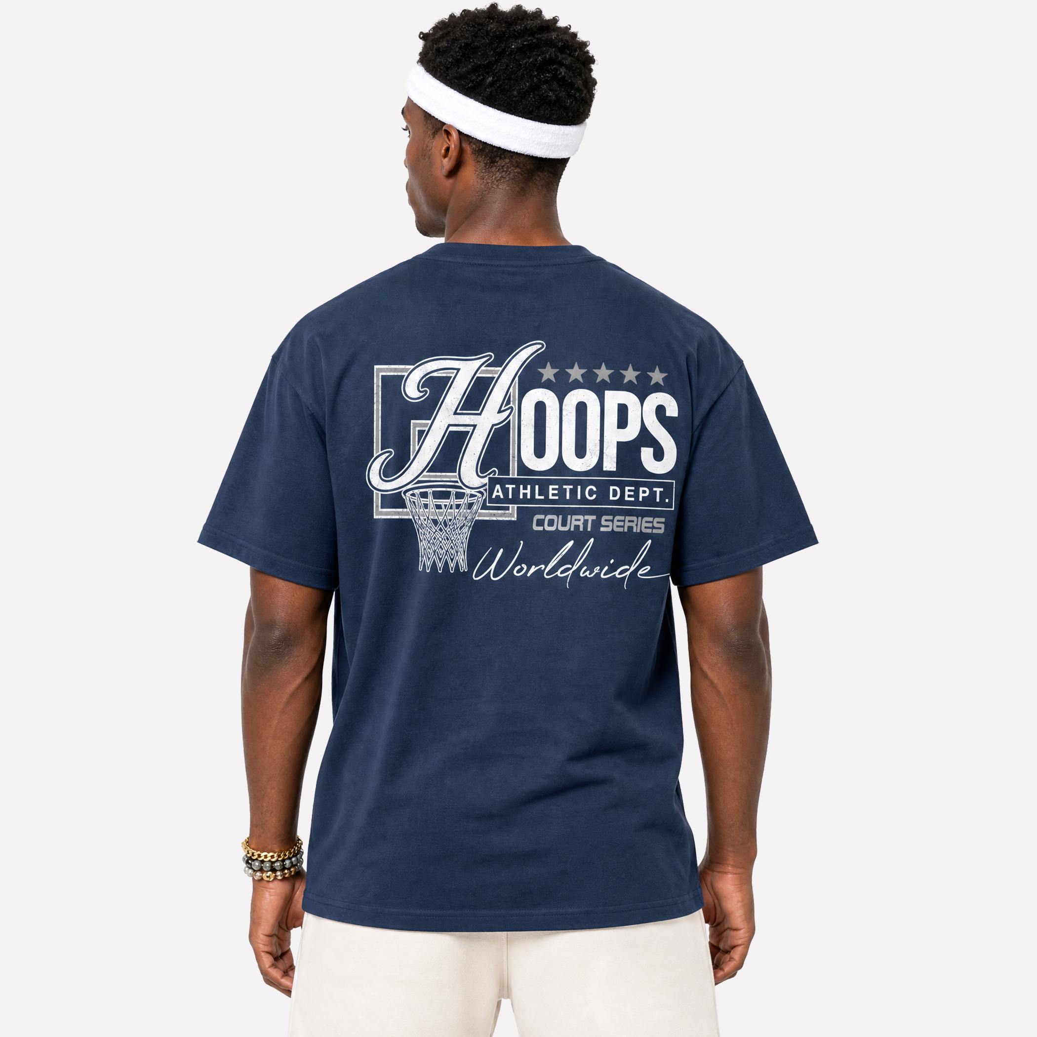 A man viewed from the back wearing a navy blue t-shirt with a large white graphic on the back that reads 'HOOPS ATHLETIC DEPT. COURT SERIES Worldwide' and a basketball hoop illustration, paired with white pants and a white headband against a light gray background.