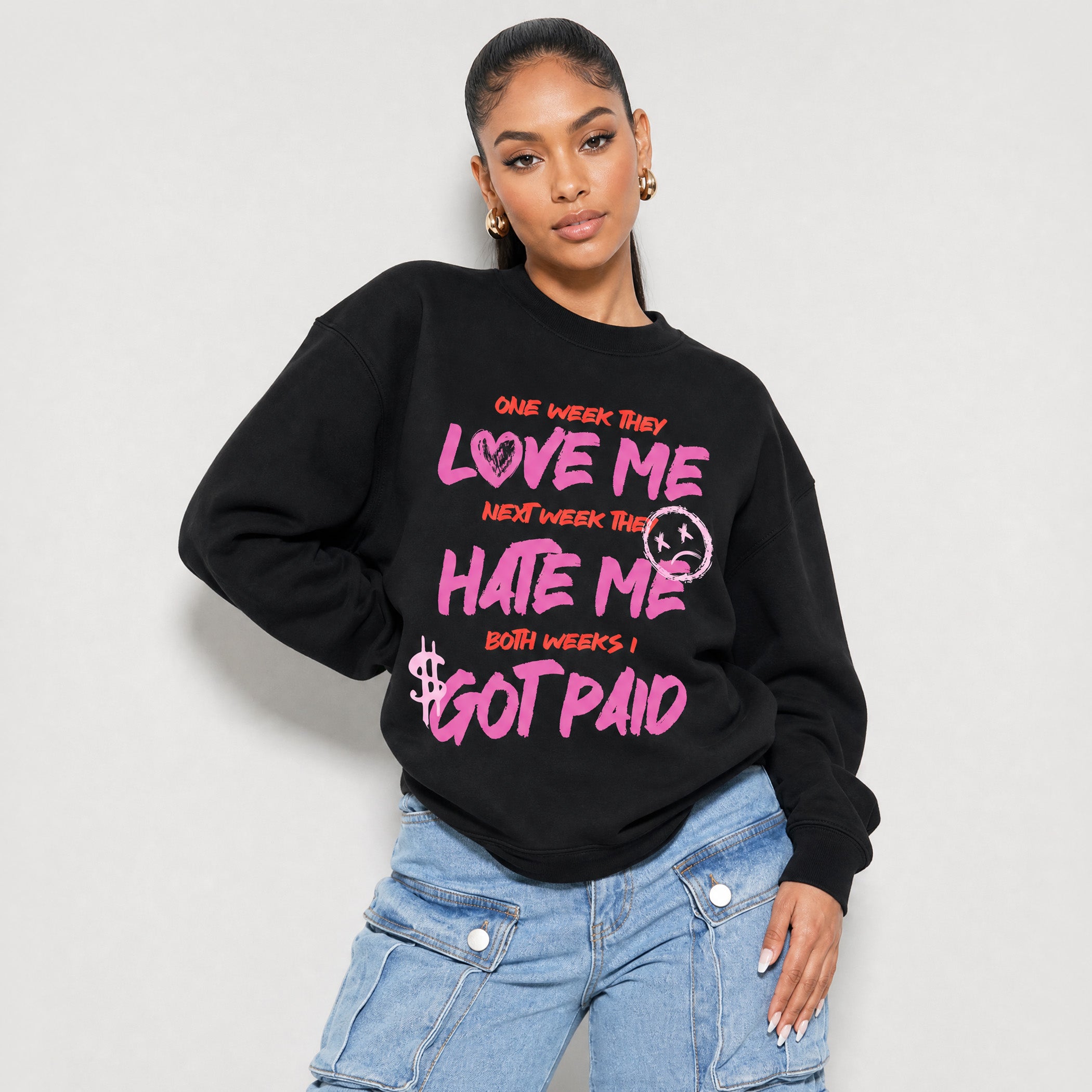 A woman modeling a black crewneck sweatshirt with a colorful graphic print that reads 'ONE WEEK THEY LOVE ME NEXT WEEK THEY HATE ME BOTH WEEKS I GOT PAID' in pink, orange, and white, paired with light blue denim cargo pants against a light gray studio background.