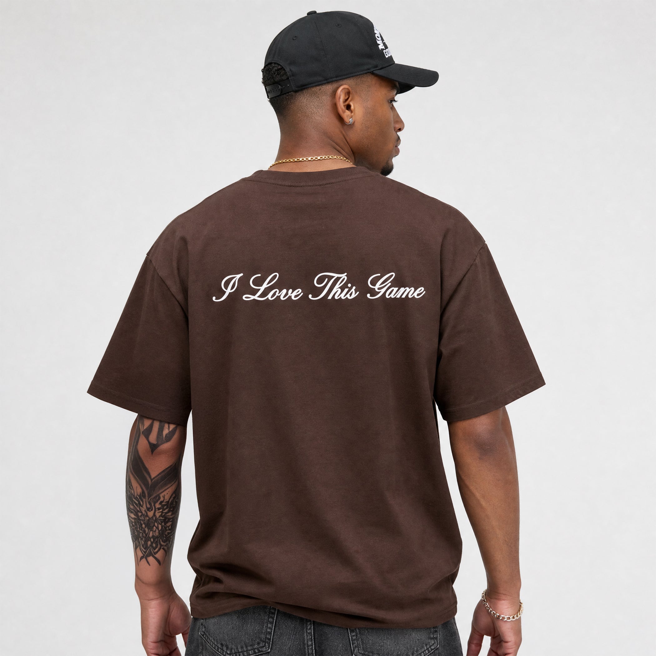 A man wearing a dark brown t-shirt with 'I Love This Game' printed in white script across the back, paired with a black baseball cap, shown from behind against a light gray background.