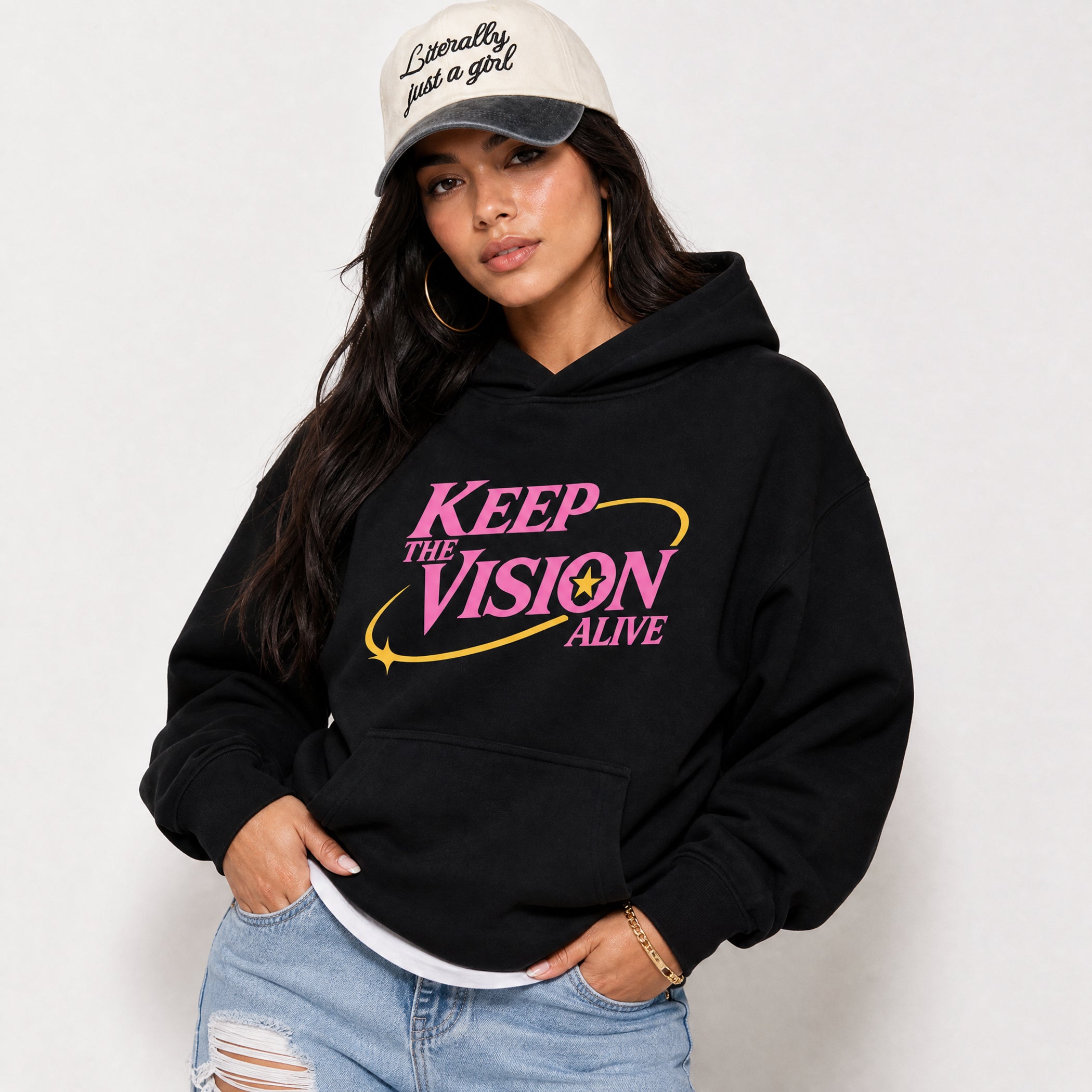 A woman wearing a black pullover hoodie with a pink and yellow graphic that reads 'KEEP THE VISION ALIVE' on the front, paired with light blue ripped jeans and a beige baseball cap with black text.