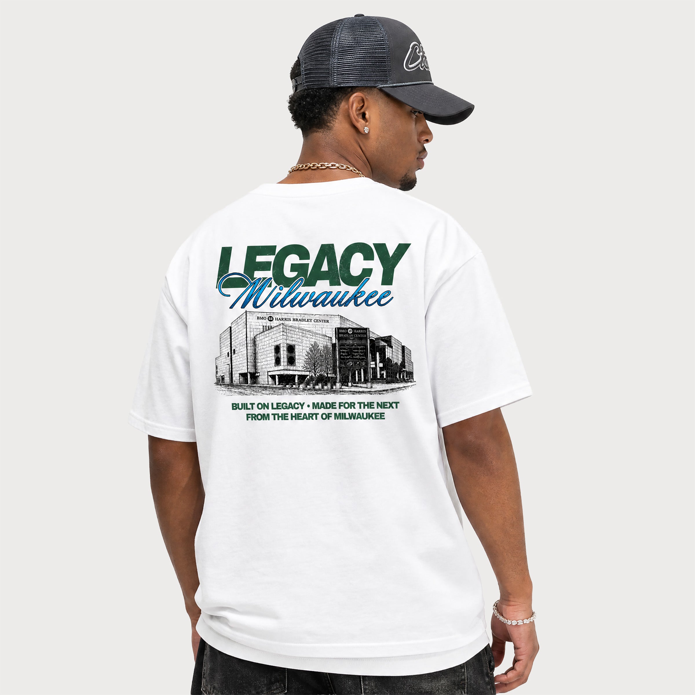 A man wearing a white graphic t-shirt from the back, featuring a green and blue 'Legacy Milwaukee' print with a black-and-white building illustration. He is also wearing a dark gray trucker cap and dark jeans, photographed against a solid light gray background.