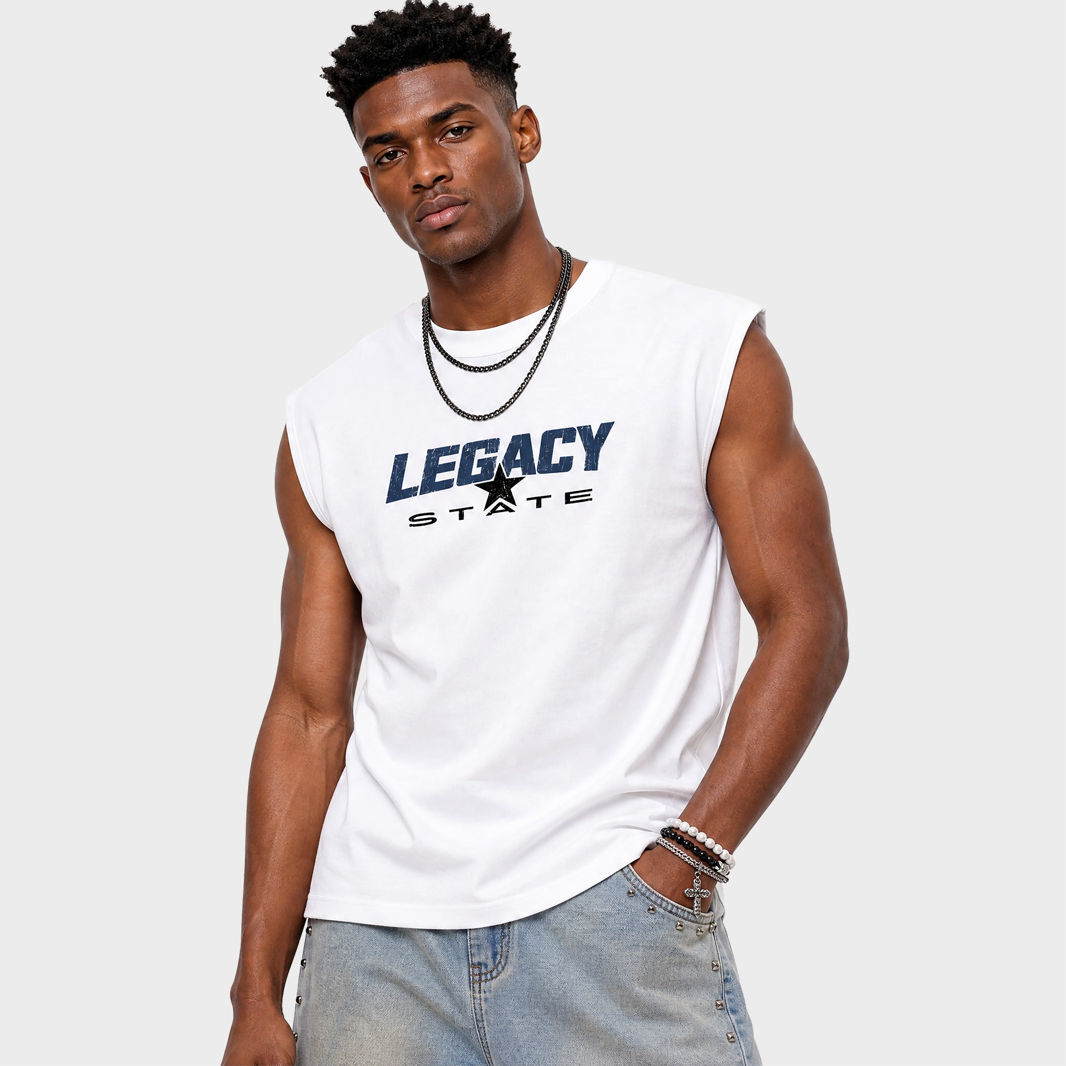 A man wearing a white sleeveless muscle t-shirt with 'LEGACY STATE' printed in navy blue and black on the chest, paired with light-wash jeans, against a light gray background.