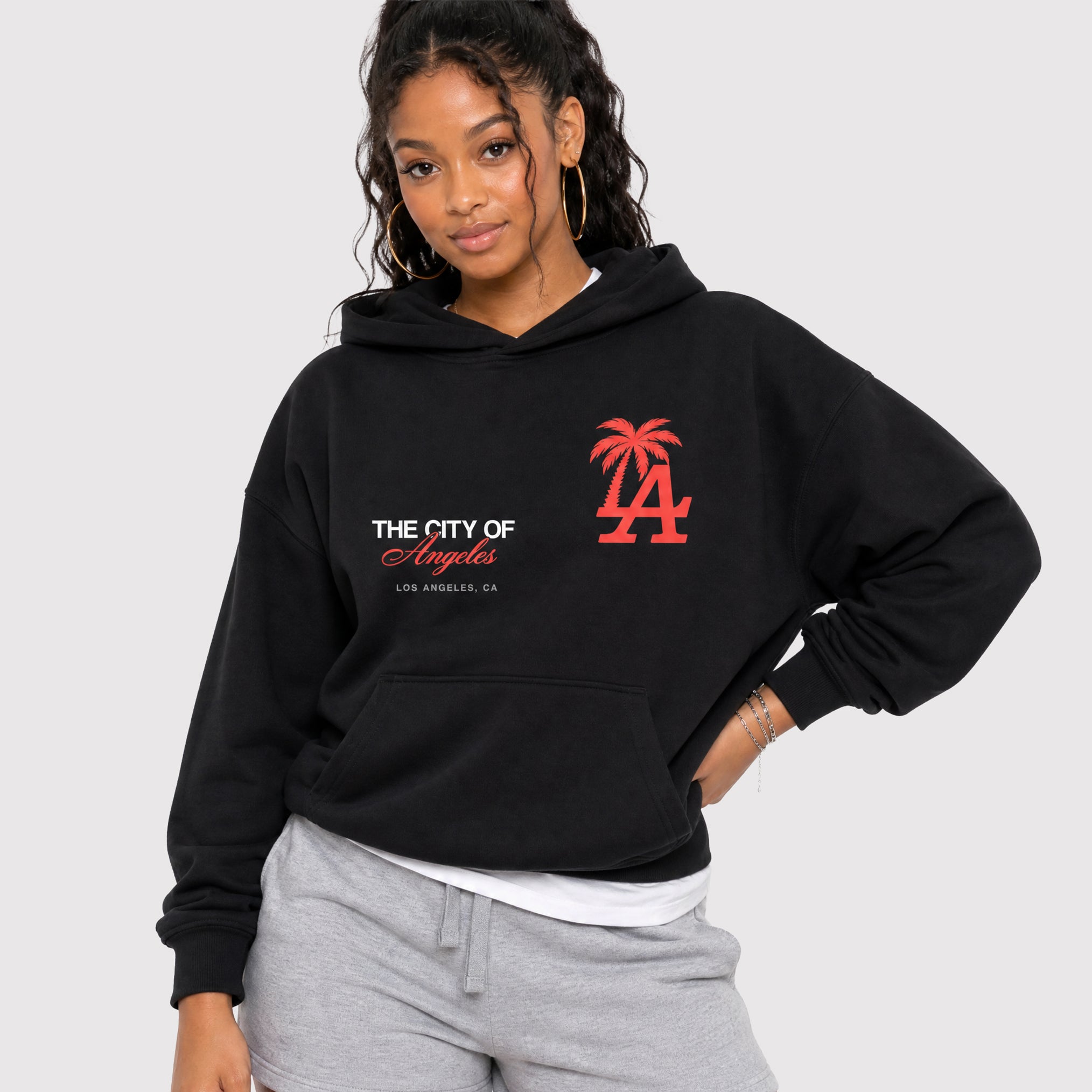 A woman wearing a black pullover hoodie with red and white Los Angeles graphics, paired with gray sweatpants, against a light gray studio background.