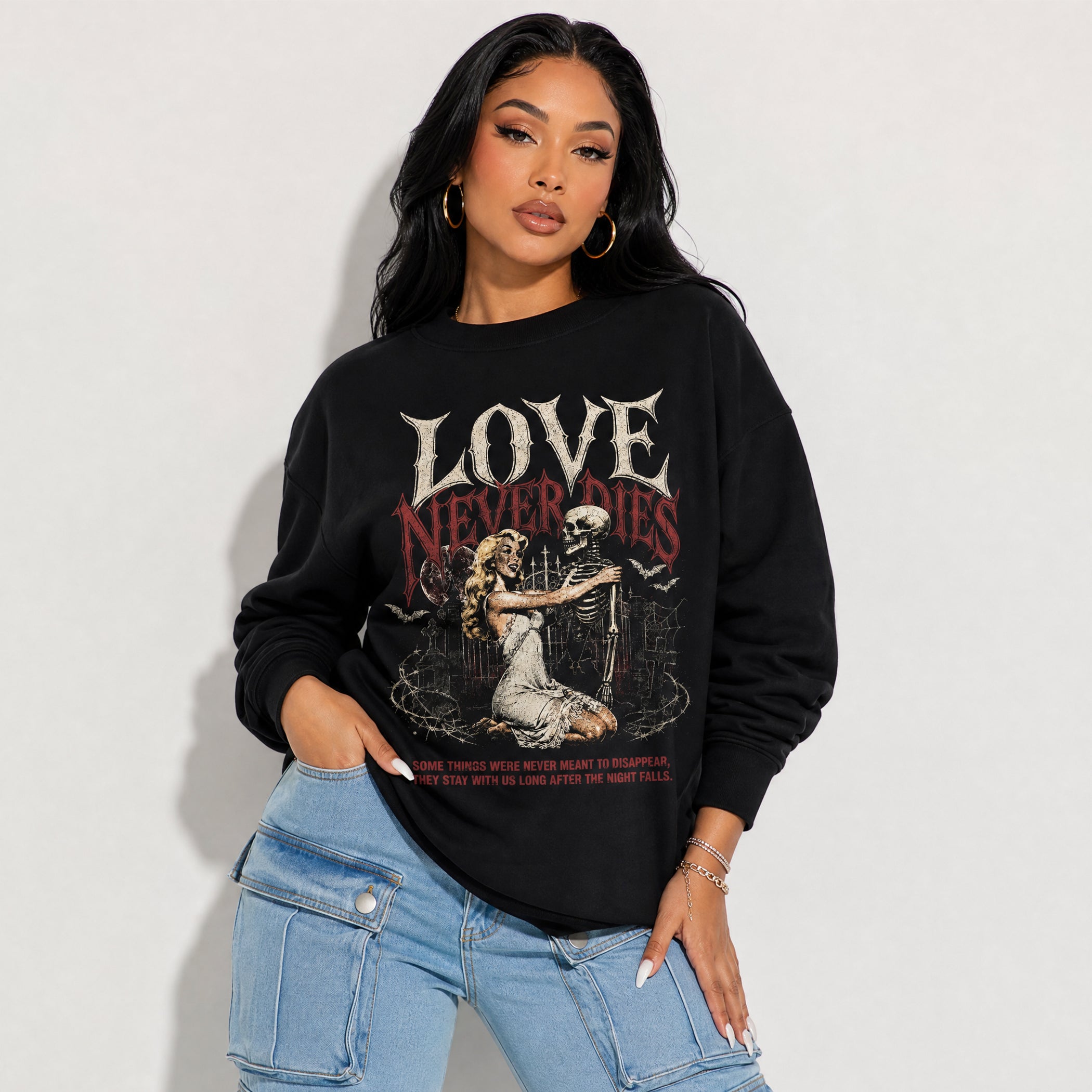 A woman wearing a black crewneck sweatshirt with a graphic print that reads 'LOVE NEVER Dies' above an illustration of a woman and skeleton, paired with light blue jeans against a plain white background.