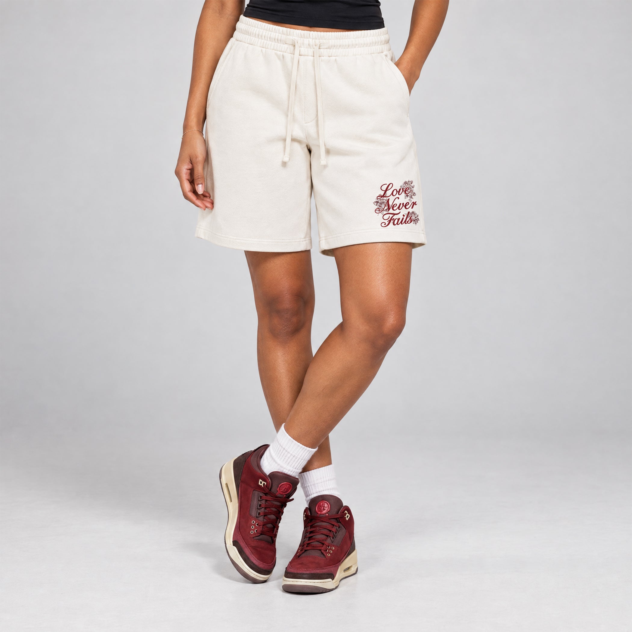 A model wearing off-white sweat shorts with a drawstring waist and 'Let Me Never Fall' embroidered on the left leg, styled with white socks and burgundy sneakers against a light gray studio background.